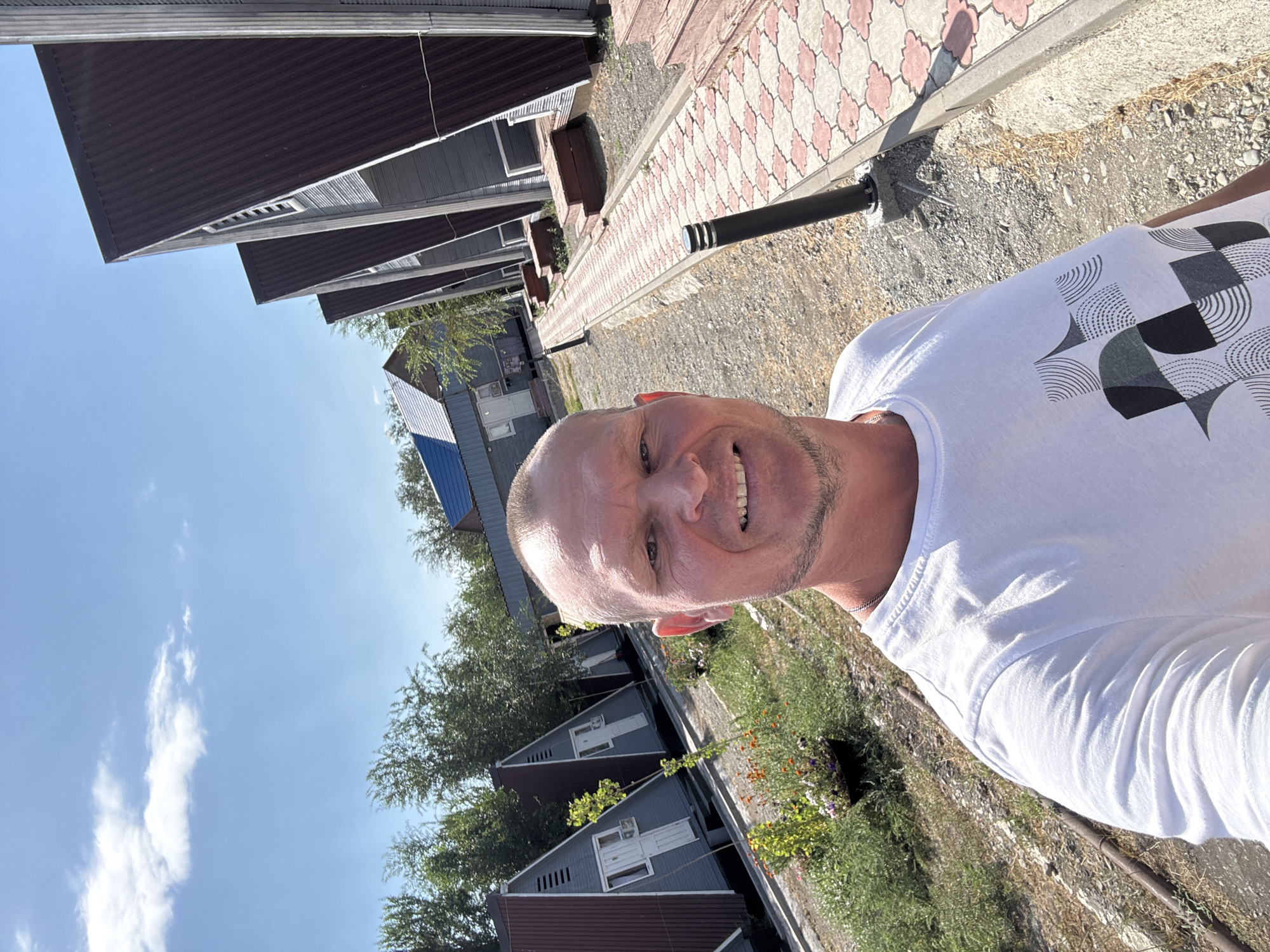 A man in a white t-shirt takes a selfie outdoors in what appears to be a resort or holiday village in Zharbulak, Kazakhstan. He is smiling, and his head and shoulders fill the foreground of the image. Behind him, a row of A-frame cabins with dark brown, corrugated metal roofs and light grey siding stretches into the distance. The cabins are set against a backdrop of green trees and a clear blue sky with scattered white clouds. A paved walkway with a red and white patterned design runs along the side of the cabins, with a black lamppost on it. There are patches of dry grass and some small flowering plants to the left of the walkway. The overall impression is a sunny day in a vacation setting.