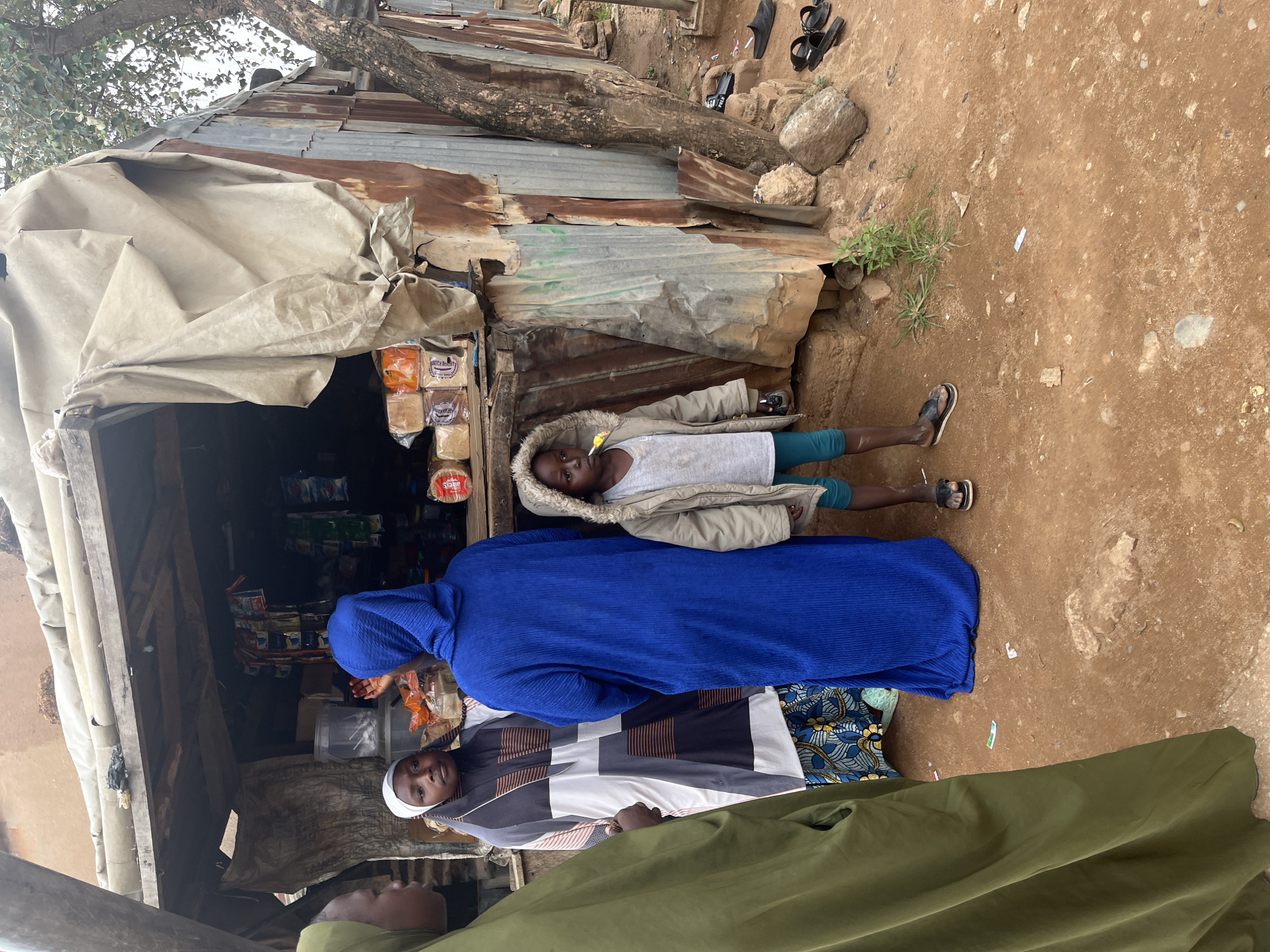 The scene is an outdoor area in Jos, Nigeria, appearing to be a small, informal marketplace or roadside vendor location. The ground is dirt and there is a building made of rusted corrugated metal. A child lies on the ground outside of a small shop. A woman in a long blue robe is standing next to another woman wearing a white head covering and a geometric-patterned tunic. Part of a person wearing a dark green garment is visible in the foreground. The shop behind them has shelves filled with various products, including packaged bread.