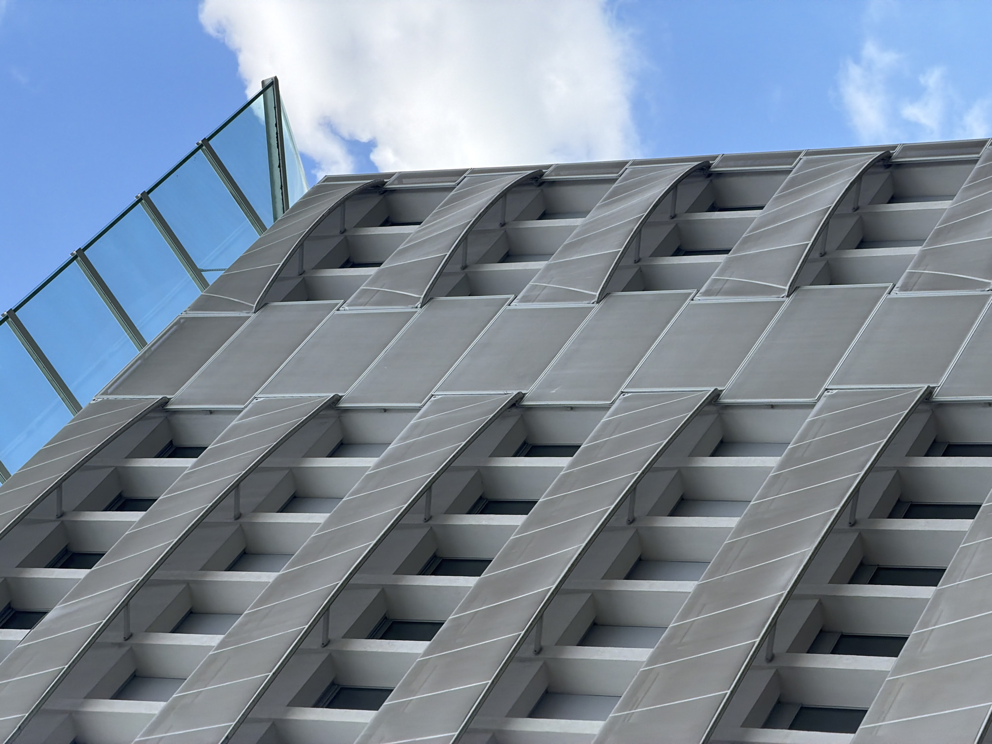 The image shows the exterior of a modern building in Vélizy-Villacoublay, France, under a blue sky with scattered clouds. The building features a facade with repeating rectangular windows, each recessed within a white frame. Horizontal white panels with diagonal lines run along the facade, creating a textured appearance. A glass canopy, supported by metal beams, extends from the top of the building. The architectural style suggests a contemporary office or institutional building, common in business districts like Vélizy's. No people are visible in the image.