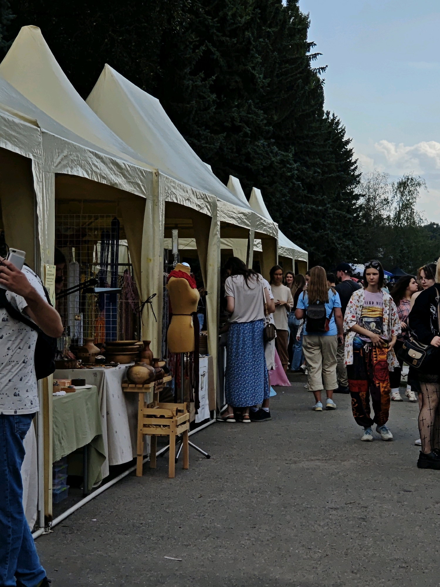 The image depicts an outdoor market scene with several white tents set up along a paved pathway. A dense line of dark green pine trees forms a backdrop. A group of people are browsing the stalls, with some standing and others walking. The stalls appear to be displaying handmade crafts and goods, including pottery, textiles, and mannequins. The sky is a pale blue with scattered white clouds. One person in the foreground is holding a smartphone, while another person on the right is wearing patterned pants and a patterned jacket. The overall atmosphere suggests a lively community event. The location is identified as Vinnytsia, Ukraine.