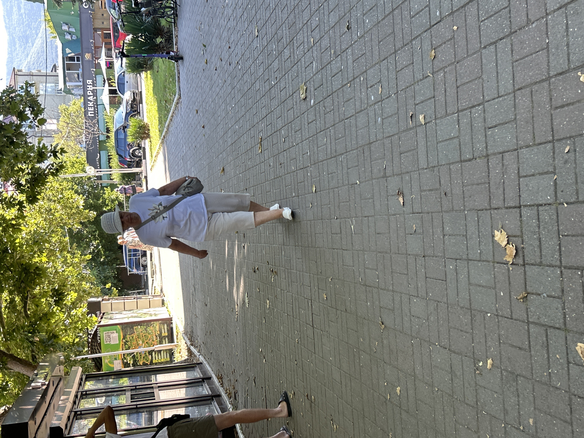 The image depicts a pedestrian walkway in Gelendzhik, Russia. A woman wearing a white t-shirt, beige shorts, and a light-colored hat is walking away from the camera. She carries a gray crossbody bag. To her left, a person in green shorts and a white t-shirt is visible, partially obscured. The walkway is paved with gray brick pavers and is lined with trees providing shade. In the background, businesses with signage, including one that reads "ПЕКАРНЯ" (Bakery), are visible. Several cars and an electric scooter are parked or in use on the street adjacent to the walkway. The environment appears to be an urban setting with commercial establishments and signs of public infrastructure.