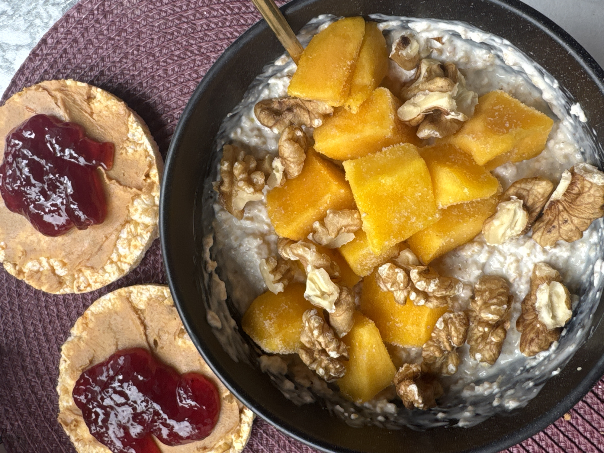 An overhead, close-up shot captures a breakfast or snack arrangement on a dark purple/brown textured placemat, likely in Khimki, Russia. The scene features a dark-colored bowl filled with a creamy, light-colored oatmeal or porridge, generously topped with bright yellow, cubed mango pieces and several scattered walnuts. A spoon with a dark handle is partially visible in the upper part of the bowl. To the left of the bowl are two round, lightly textured rice cakes. Each rice cake is spread with a layer of light brown peanut butter and topped with a glistening dollop of red fruit jam. The lighting is bright, highlighting the colors and textures of the food items. No people or visible text are present in the image.