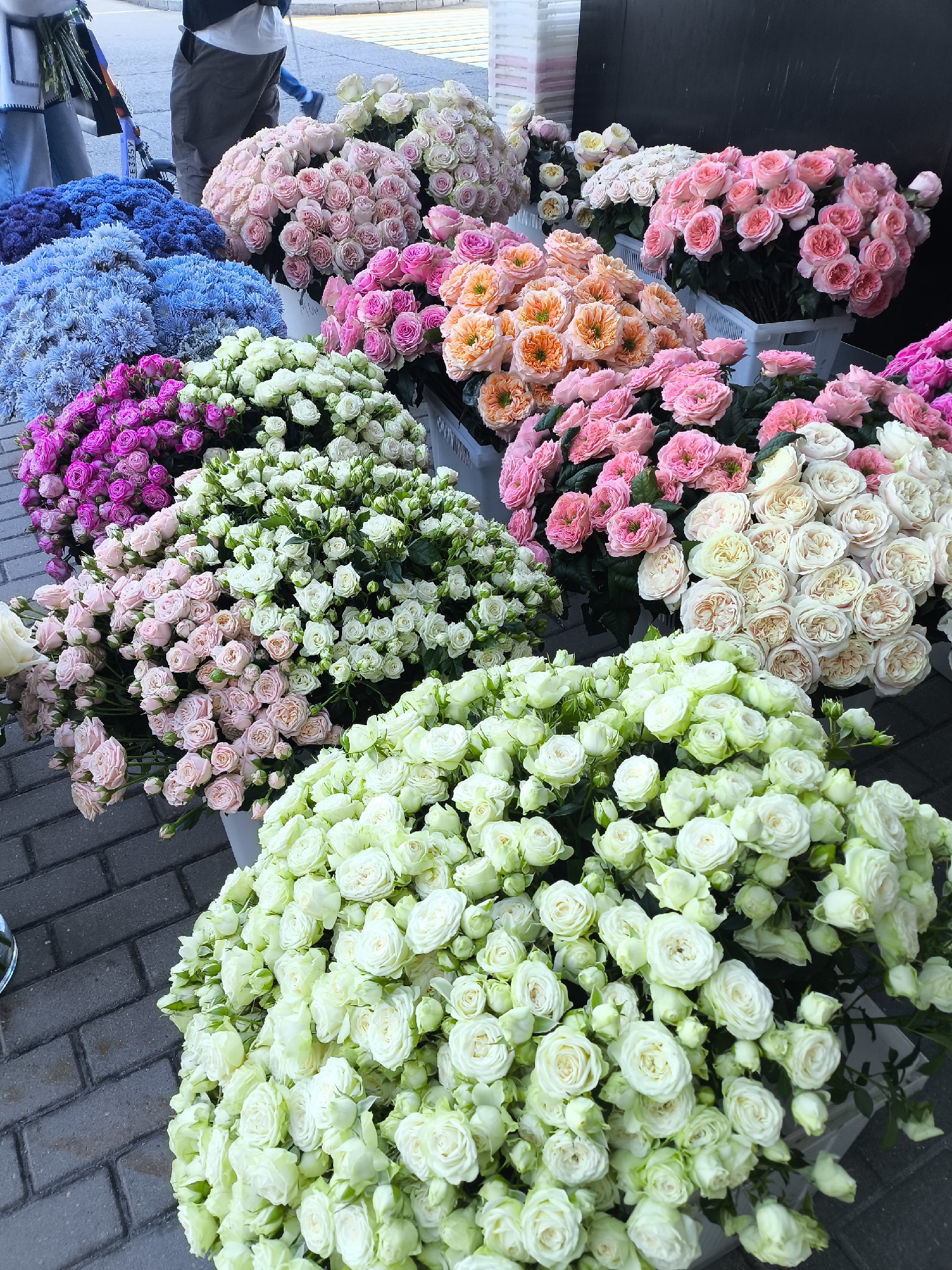 A street-level outdoor flower market stall is depicted, displaying a vibrant array of roses in various colors. The stall is situated on a brick-paved sidewalk, characteristic of urban areas. In the background, a dark, nondescript wall forms the backdrop, with some white plastic crates visible. A partial view of people in casual attire, one holding a bouquet of flowers, suggests a commercial transaction or preparation for sales. The focus is on the abundant floral display, featuring tightly packed bouquets of pink, white, blue, purple, and peach-colored roses. The lighting suggests it is daytime. The location in the city of Khimki, Russia is a detail not visually discernible from the image content.