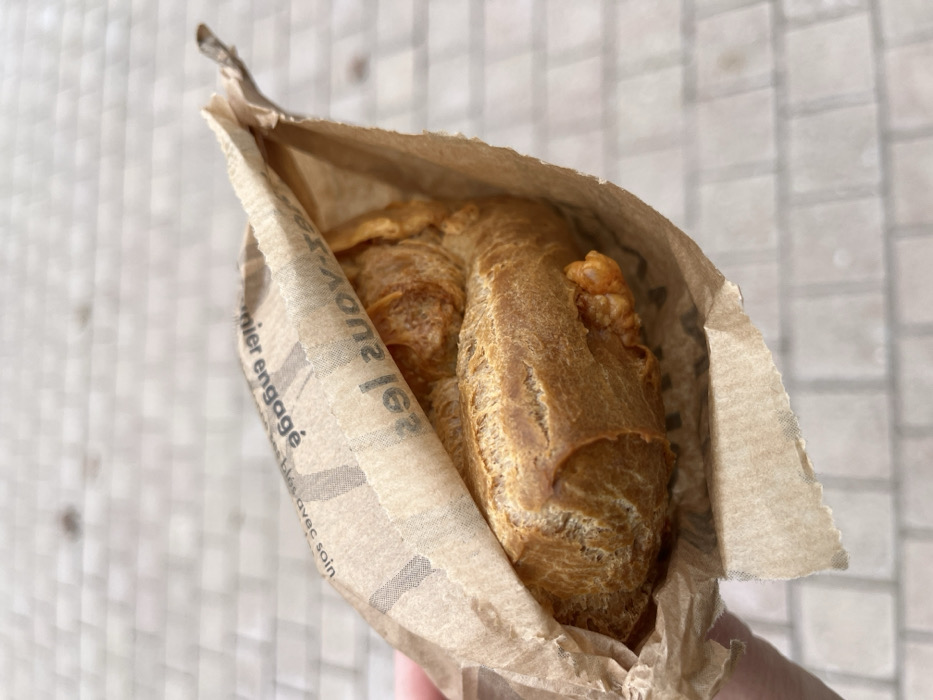 A single, elongated piece of golden-brown bread, resembling a demi-baguette or ficelle, is partially contained within a crumpled, light brown paper bag. The bag features inverted French text fragments including "nier engagé," "avec soin," and "les." A light-skinned hand is partially visible on the lower-left, gripping the bag. The bread exhibits a crusty surface, with irregular, golden-orange protuberances visible from its lower side, indicative of a filling. The background shows an out-of-focus, light-toned paved surface composed of regularly patterned rectangular blocks, consistent with an outdoor public space in Dijon, France.