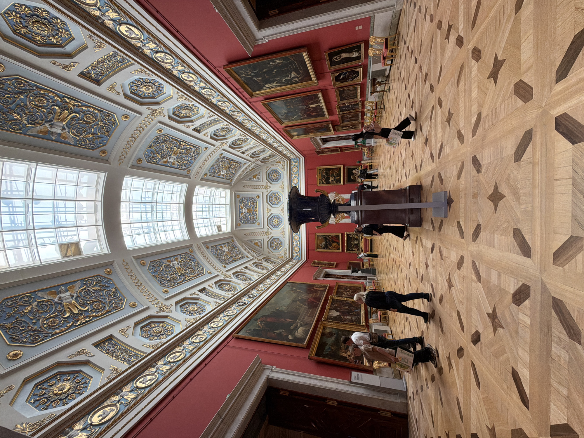 The image captures an interior view of a grand art museum, likely in Saint Petersburg, Russia, given the context. The scene is dominated by opulent architecture, with a highly decorated ceiling featuring intricate gold and blue stucco work, set against a deep red wall. Large, arched windows along the top of the room allow natural light to flood in, illuminating the space. The floor is made of intricately patterned parquet wood.
Several people are visible in the image, appearing to be museum visitors. They are casually dressed and seem to be engaged in observing the artwork on display. Some are standing and looking at paintings, while others are walking through the hall. Their postures suggest a relaxed and contemplative atmosphere, typical of a museum visit.
The walls are adorned with numerous framed paintings, suggesting a significant art collection. The paintings themselves are varied in size and style, some appearing to be old master works. In the foreground, a large, dark vase or sculpture sits on a pedestal, adding to the sense of grandeur. The overall impression is one of historical significance and artistic richness. The lighting suggests it is daytime, and the interior setting implies controlled indoor conditions, irrespective of external weather. No visible text is discernible within the image.