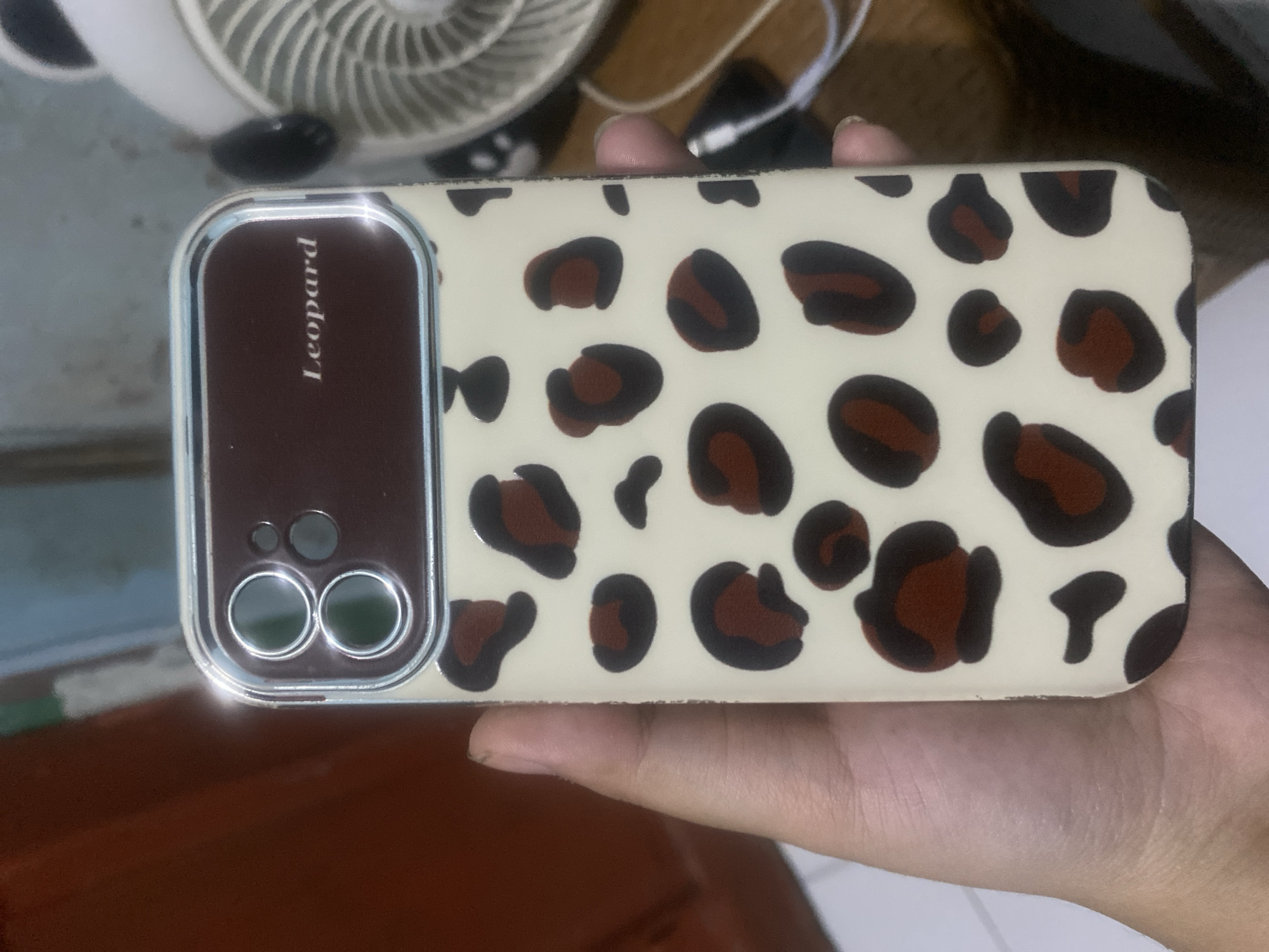 The image shows a close-up of a person's hand holding a beige smartphone case with a leopard print pattern. The case has a dark brown section at the top, framing the camera lenses, and includes the text "Leopard" in white lettering. The background is blurred, but appears to be an indoor setting with wooden furniture and a white fan. There are no discernible landmarks or indicators that place this scene specifically in Kalitinggar, Indonesia.