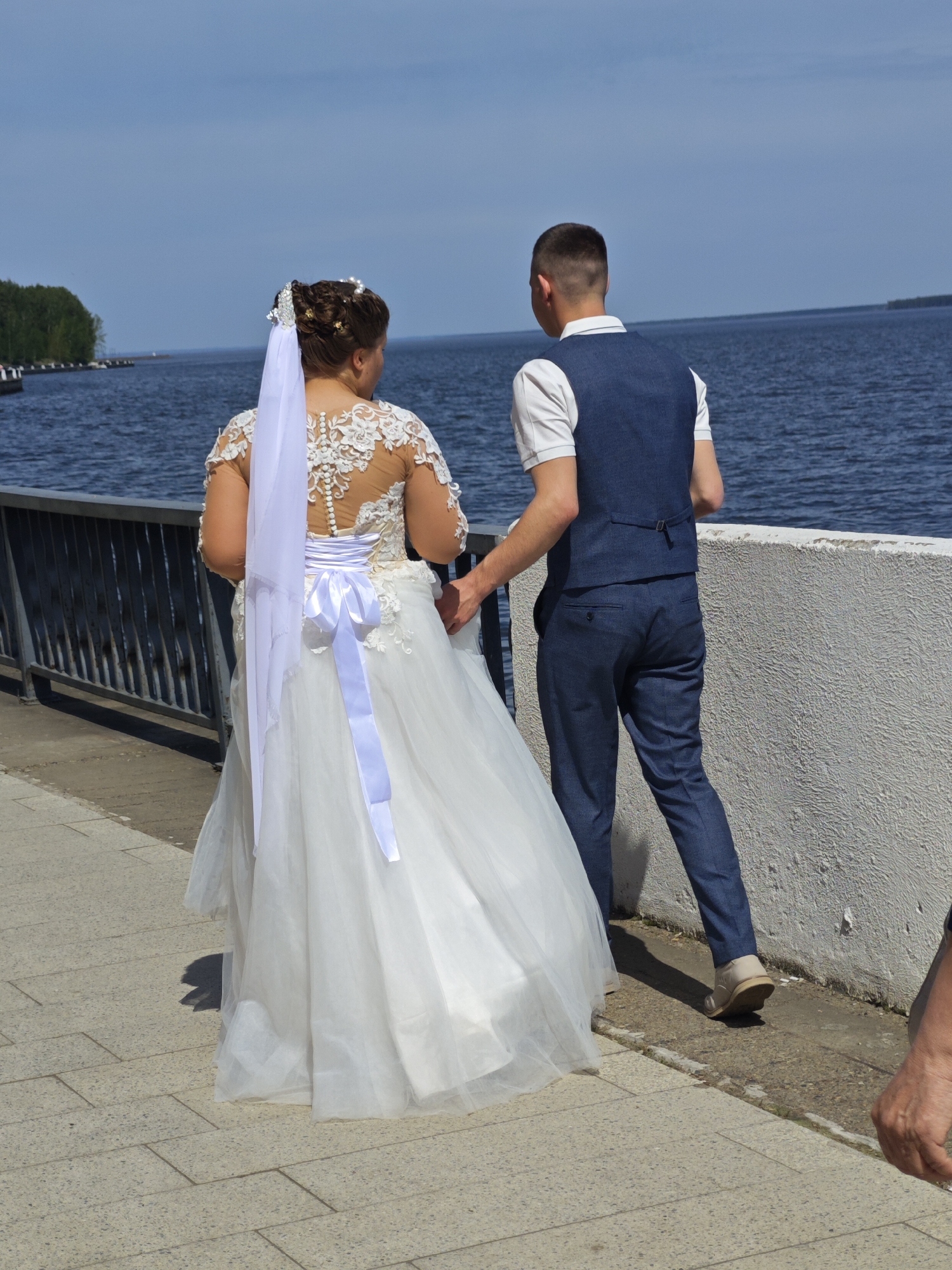 A couple, presumably newlyweds, is walking along a promenade. The bride is wearing a white wedding dress with lace detailing on the back and a long white veil secured to her hair. The dress has a large white satin ribbon tied in a bow at the waist. The groom is wearing a blue vest over a white short-sleeved shirt and matching blue pants. He has his hand gently placed on the bride's arm. They are walking away from the viewer, towards a body of water. A metal railing runs along one side of the promenade, with a low white wall on the other. In the background, a forested area is visible across the water, with some structures along the shoreline. The promenade is paved with light grey paving stones. A portion of another person's arm is visible in the lower right corner of the image. The scene appears to be in Chaikovsky, Russia, based on the location and the style of the architecture and landscape.