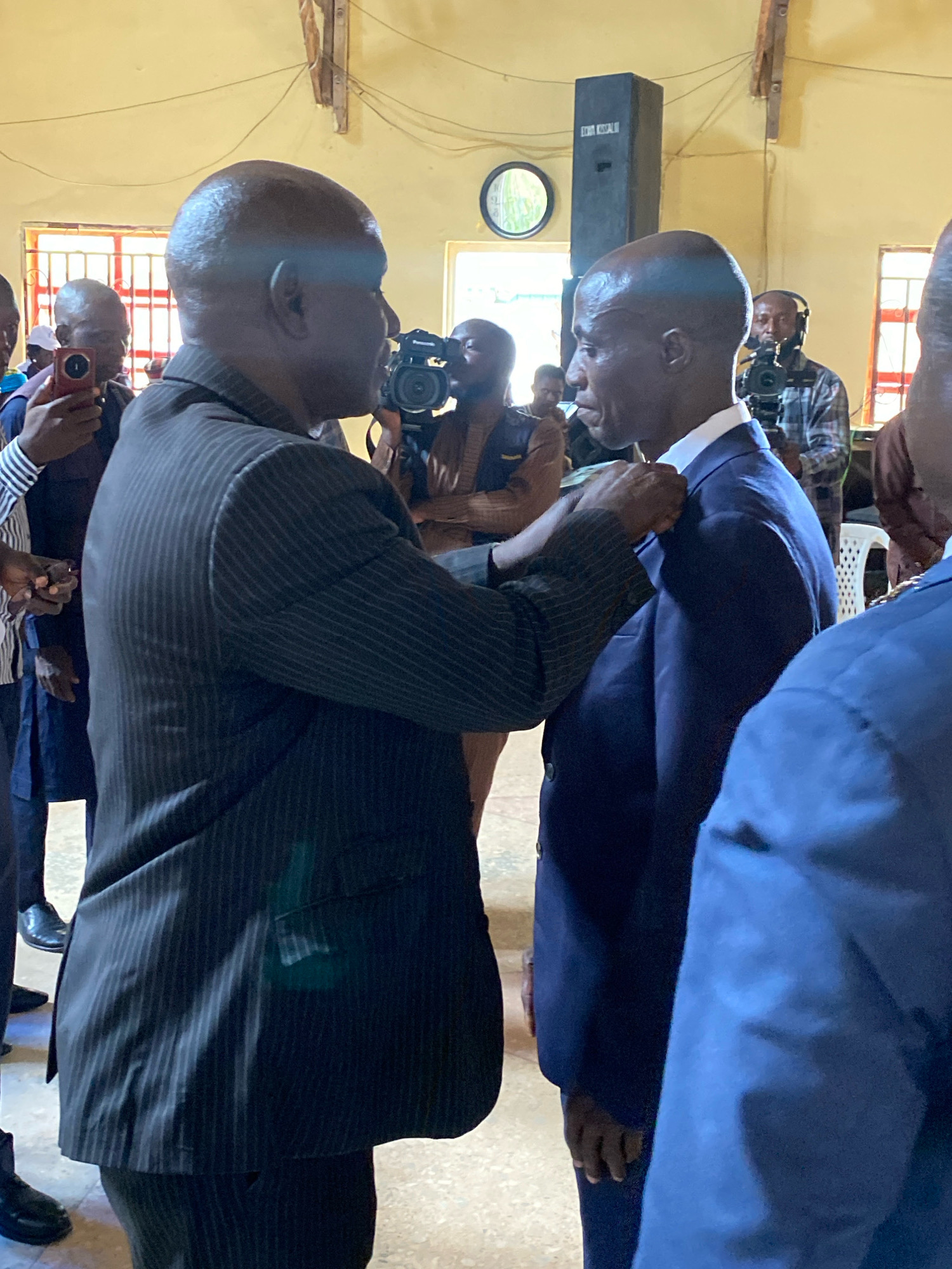 In an indoor setting, likely a community hall in Ohit, Nigeria, two men are the central focus of the image. The man on the left, wearing a pinstripe suit jacket and trousers, is in the process of pinning a lapel pin onto the suit jacket of the man on the right. The man on the right is wearing a dark blue suit jacket and shirt. Several other individuals are visible in the background, some holding cameras and mobile phones, suggesting an event or ceremony is taking place. The room appears to be sparsely furnished, with visible electrical conduits and a speaker system. A round clock is visible on the wall in the background.