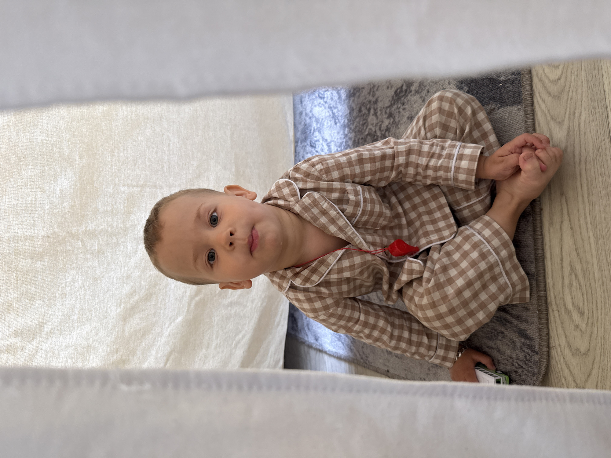 A child wearing brown and white plaid pajamas is sitting cross-legged on a gray and white rug. The child has fair skin, short hair, and is looking directly at the camera with a slight smile. A red whistle is visible around the child's neck. A white cloth or curtain is partially visible in the foreground. The rug is on a light-colored wooden floor. The location appears to be indoors, possibly in a home. No location in Rusanovo, Russia, can be determined from this image.