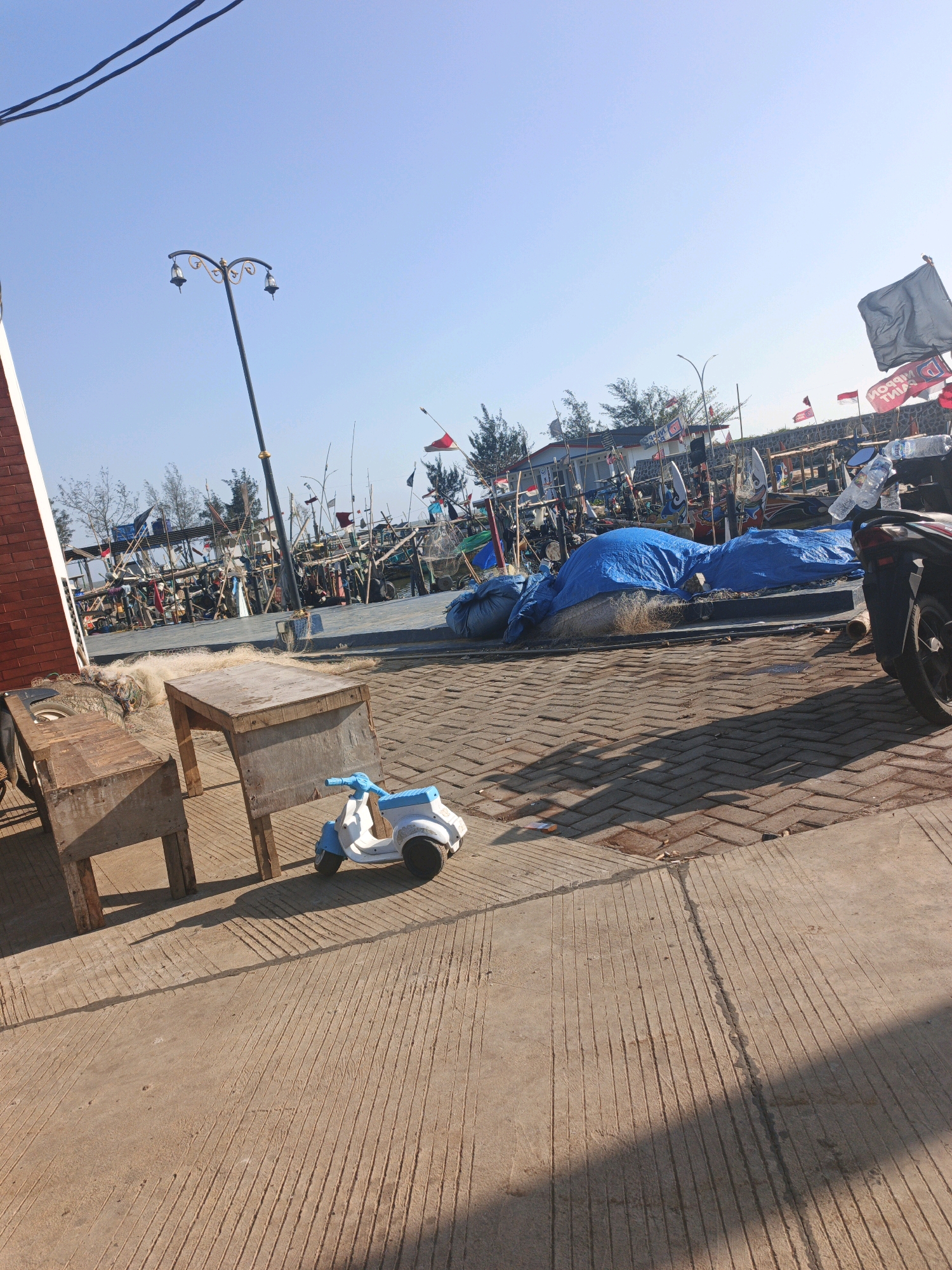 In Unknown, Indonesia, a sunny outdoor scene depicts a paved area with a prominent shadow cast by a wooden table and chairs. In the foreground, a white and blue toy scooter sits on the pavement. Behind the table and chairs, fishing nets are visible. Further in the background, a street lamp stands tall against a clear blue sky. Numerous fishing boats are moored in the distance, adorned with many flags. A motorcycle is parked on the right side of the frame, with a plastic bottle of water on its handlebars. The environment appears to be a coastal area, possibly a harbor or pier, given the presence of fishing boats and nets.