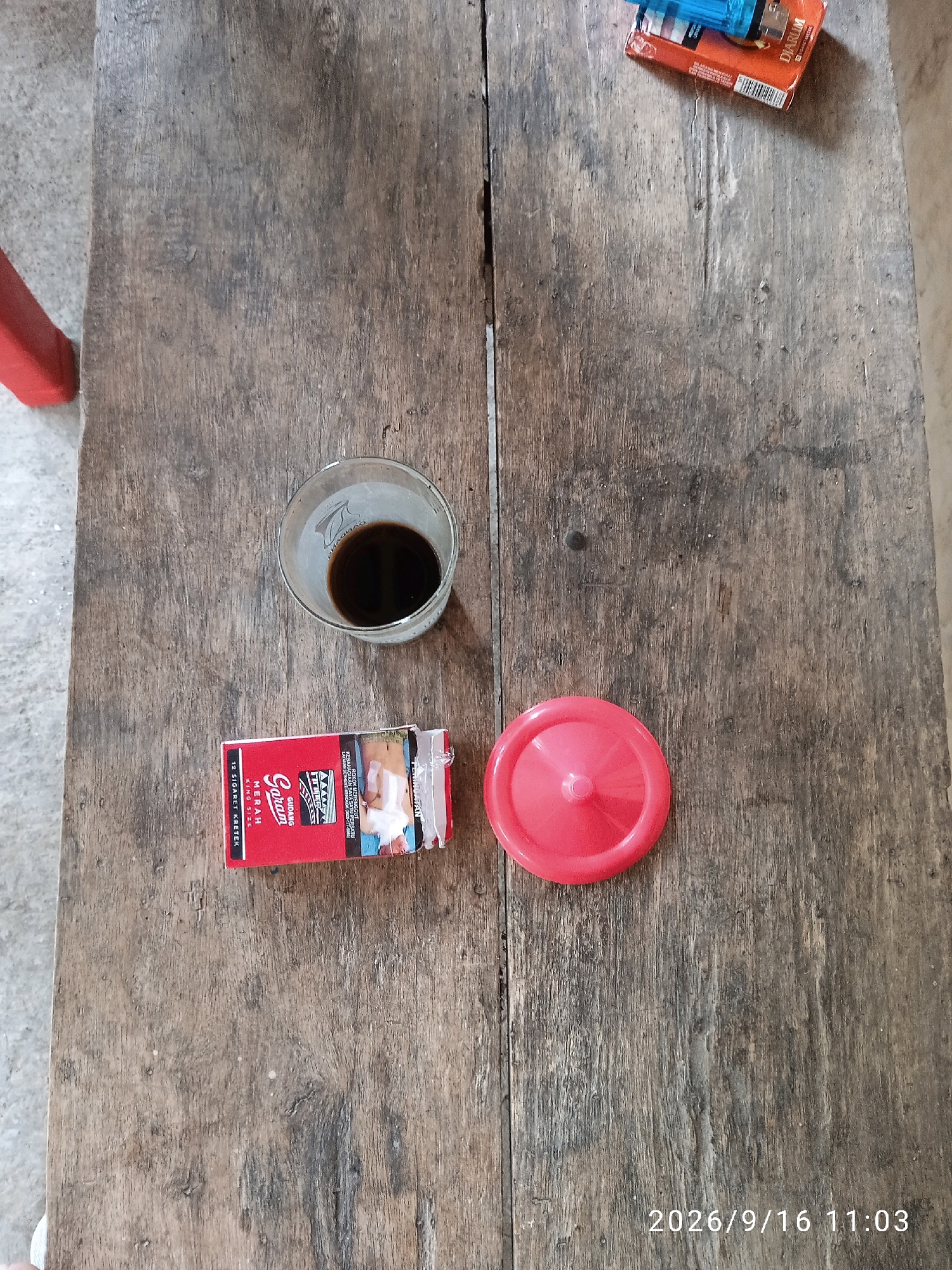 A still life photograph shows a rustic wooden table with various objects on it. In the center, a glass of dark liquid, likely coffee, sits next to an open pack of red cigarettes labeled "Gudang Garam Merah KING SIZE." To the right of the cigarettes is a bright red plastic lid. In the upper right corner, a blue and orange lighter rests on the table, partially obscuring another orange pack. The table surface is weathered and shows visible wood grain. In the lower right corner, a timestamp indicates "2026/9/16 11:03." A red chair leg is visible on the left edge of the image. The scene appears to be set in an outdoor or semi-outdoor environment with a muted grey background. The overall impression is one of a simple, everyday moment.