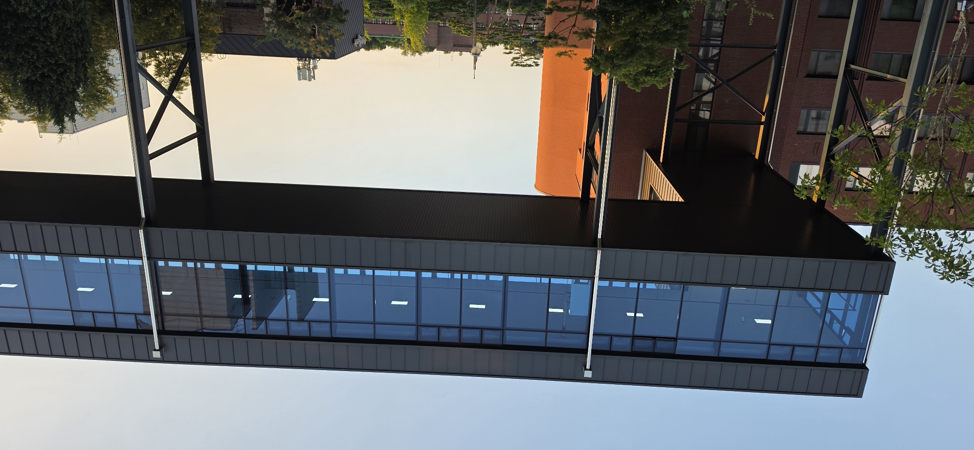 A modern skybridge with a dark gray metal exterior and a glass facade is suspended between two buildings. The skybridge has a row of rectangular windows, each containing a white light fixture. Below the skybridge, a brick building with a curved orange facade is visible on the right, and a darker brick building with several windows is on the left. The scene is set against a pale blue sky with a gradient of light yellow towards the horizon, suggesting either sunrise or sunset. Trees are scattered throughout the landscape, with a leafy tree prominently positioned in the foreground on the left. In the distance, other buildings, including a taller white structure, and more trees are visible, hinting at an urban or campus environment. The image was taken in Cheongju-si, South Korea.