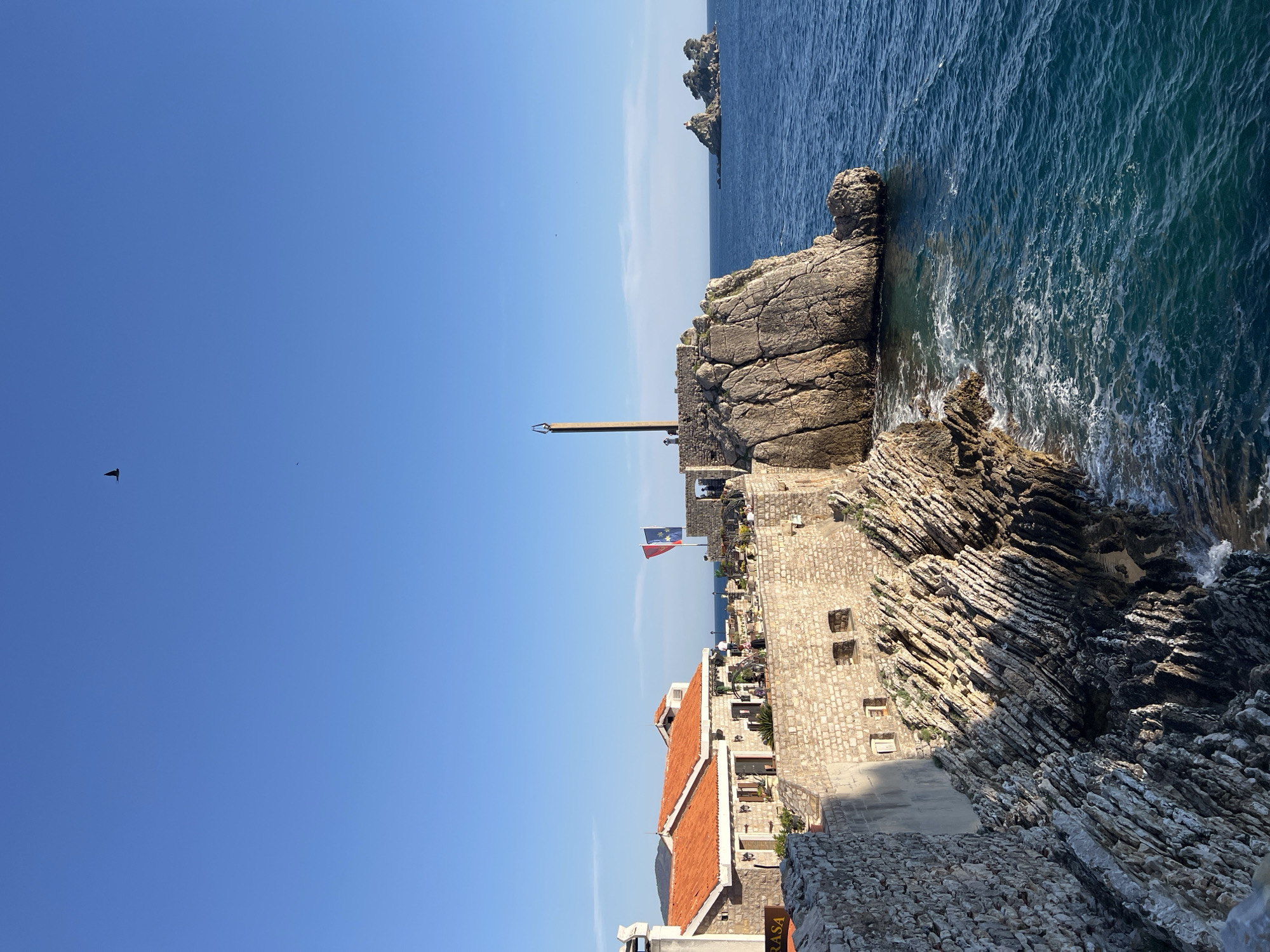 The image depicts a coastal scene in Petrovac centar, Montenegro. The foreground features weathered stone buildings with red-tiled roofs and stone walls, partially covered in moss and vegetation. A flag, possibly the flag of Montenegro, is visible atop a building. Further out, a rocky promontory extends into the Adriatic Sea. The sea is a deep blue, with visible wave action against the rocks. In the distance, smaller rocky islands are seen. A clear blue sky covers most of the frame, with a few birds visible. A metal pole stands atop the buildings near the edge of the rocks.