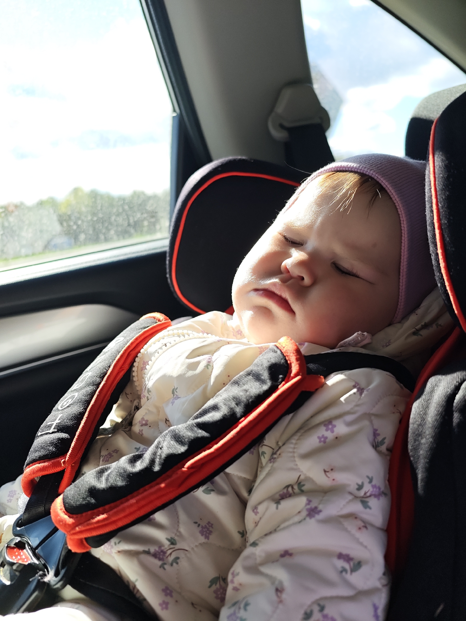 A baby wearing a pink beanie and a white floral jacket is asleep in a black and orange car seat. The baby is secured with the car seat harness. Light is entering the car through the window on the left, revealing an out-of-focus landscape with trees. The photograph was taken inside a car, likely in Chaikovsky, Russia.