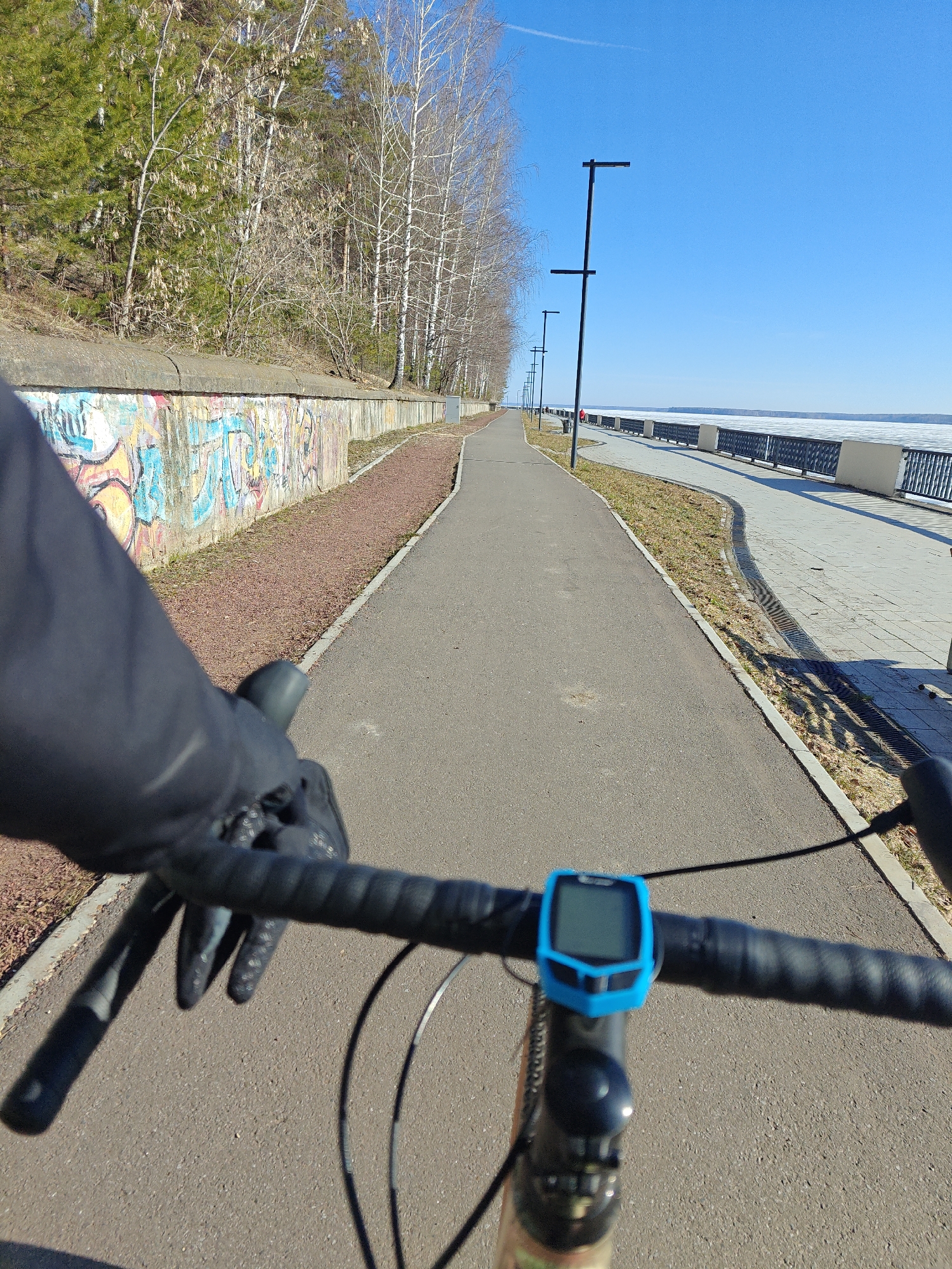 The image presents a first-person perspective from a cyclist riding on a paved path, likely in the city of Chaikovsky, Russia, based on the presence of the Kama Reservoir. The cyclist's gloved left hand is visible on the handlebars, with a blue bicycle computer mounted in the center. To the left of the path is a low concrete wall covered in graffiti, with trees and vegetation behind it. To the right is the Kama Reservoir with remnants of ice still visible, alongside a paved walkway with street lamps and a metal railing. The sky is clear and blue. The environment suggests early spring, with trees yet to fully leaf out. The action is someone riding a bicycle on a pathway along the reservoir in Chaikovsky.