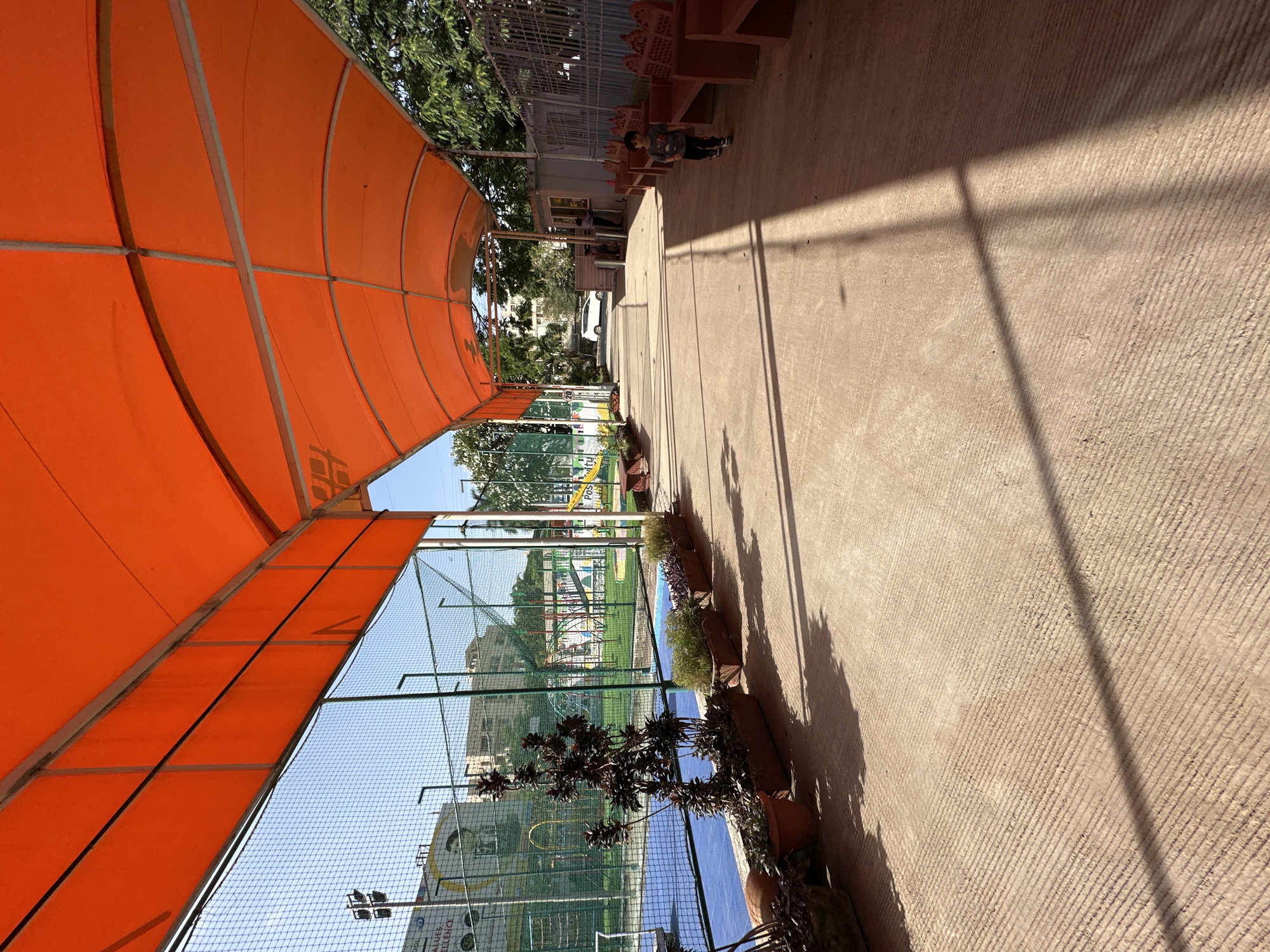 The image depicts an outdoor area in Hinjawadi, India. A structure with an orange canopy covers the left side of the frame. To the right of the canopy is a concrete pathway, marked by sunlight and shadows. Adjacent to the pathway are potted plants. Beyond a chain-link fence, a playground is visible. In the background, there are buildings and trees. Further down the pathway, a child and adults are walking. Wooden benches are on the right of the path next to the adult and child.