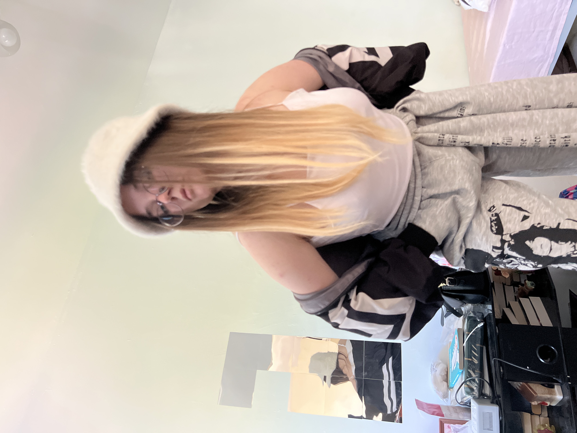 A young woman stands indoors wearing a white bucket hat, glasses, a white t-shirt, a grey hoodie with lettering, and a black and white jacket. The location could be a bedroom based on the bed visible in the upper right corner. The background includes a light green wall, a mirror consisting of several squares, and a cluttered black desk with various objects including a black purse, a charging cable, and other personal items. The exact location within Pushchino, Russia, cannot be determined from the given image.