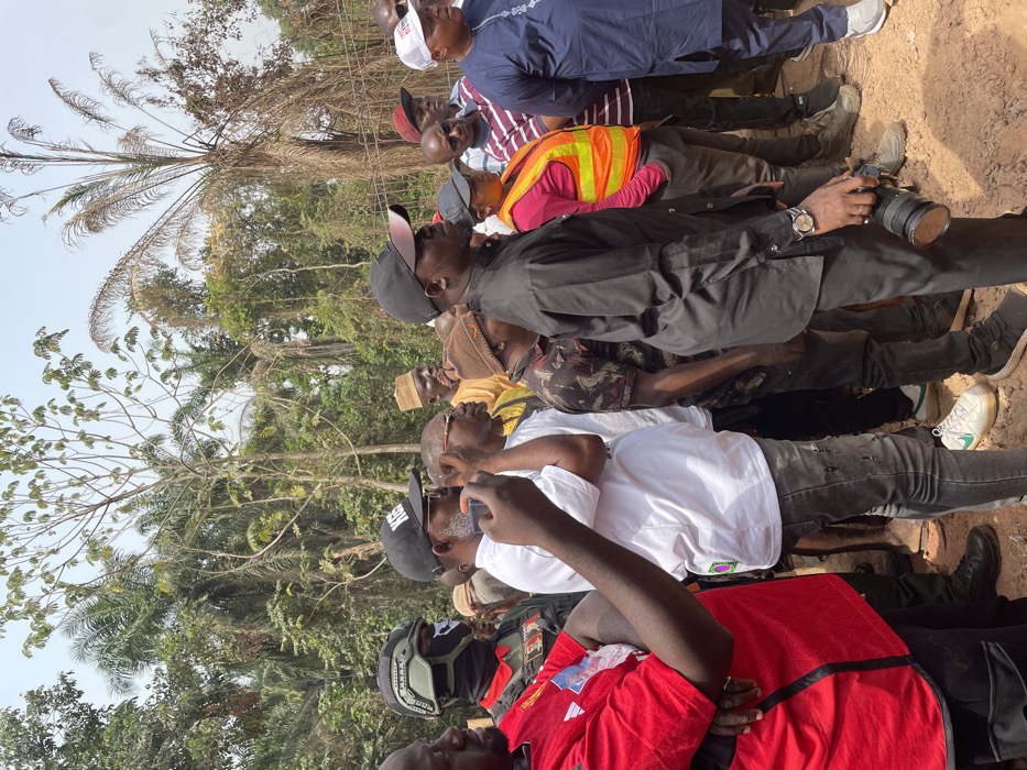 A group of approximately ten men are gathered outdoors under bright daylight in what appears to be a rural or semi-rural area in Aloko, Nigeria. The image captures them from a low angle, standing on packed dirt or sand, with dense green foliage and palm trees forming the background. The sky is clear, indicating sunny weather.
The men are dressed in various casual attire. Notable clothing includes a man wearing a bright orange safety vest over a pink/red shirt, another in a white shirt, one in a black shirt and black cap, and another in a blue long-sleeved top. Several men are wearing caps; one black cap has "SN" or "SIT" visible, and a white cap in the background shows partial text like "C_MP_SS".
Many of the men are looking upwards or forward, suggesting they are observing an event or object outside the camera's frame. One man in a black shirt is holding a professional camera with a large lens, pointing it forward. Another man in a white shirt is holding a smartphone, seemingly recording or taking a picture. The collective gathering, coupled with the presence of cameras and a safety vest, suggests they might be attending an inspection, a community event, or documenting a happening in the area. No strong emotions are clearly discernible; most appear focused on what they are observing.