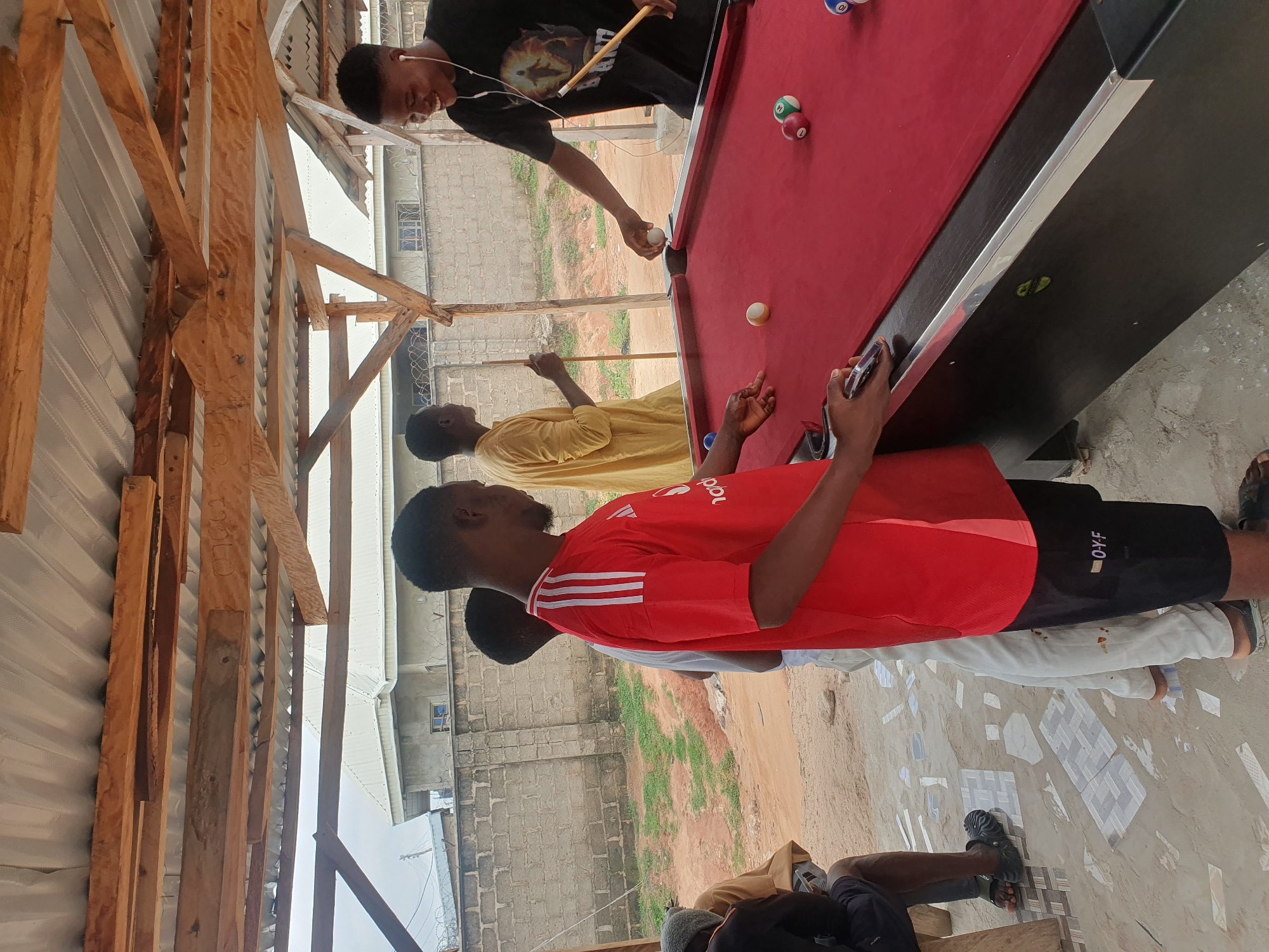 The image shows a group of young men playing billiards outdoors in what appears to be a casual setting, possibly a backyard or a makeshift entertainment area. The scene is dominated by a red billiard table with several balls visible, including a cue ball and numbered balls. Two men are actively engaged in the game. One man, wearing a black t-shirt and earphones, is holding a cue stick and looking at the table, seemingly focused on his shot. He has a slight smile, suggesting he's enjoying the game. The other man, wearing a red t-shirt with white stripes on the sleeves and black shorts, is looking towards the table, possibly waiting for his turn or observing the ongoing play.
In the background, there are structures made of corrugated metal and wooden beams, suggesting a partly constructed or open-air shelter. Beyond this, a brick wall and some vegetation are visible, hinting at a semi-urban or rural environment. The ground is a mix of dirt and some tiled or paved sections with a checkered pattern. The lighting suggests it's daytime, with natural sunlight illuminating the scene. The overall mood seems relaxed and social, with the individuals engrossed in their game. The location context provided indicates Lokoja, Nigeria. No specific time of day or weather conditions can be definitively determined, though the brightness suggests daylight. There is some text visible on the black shorts of the man in the red t-shirt which appears to read "OY-F".