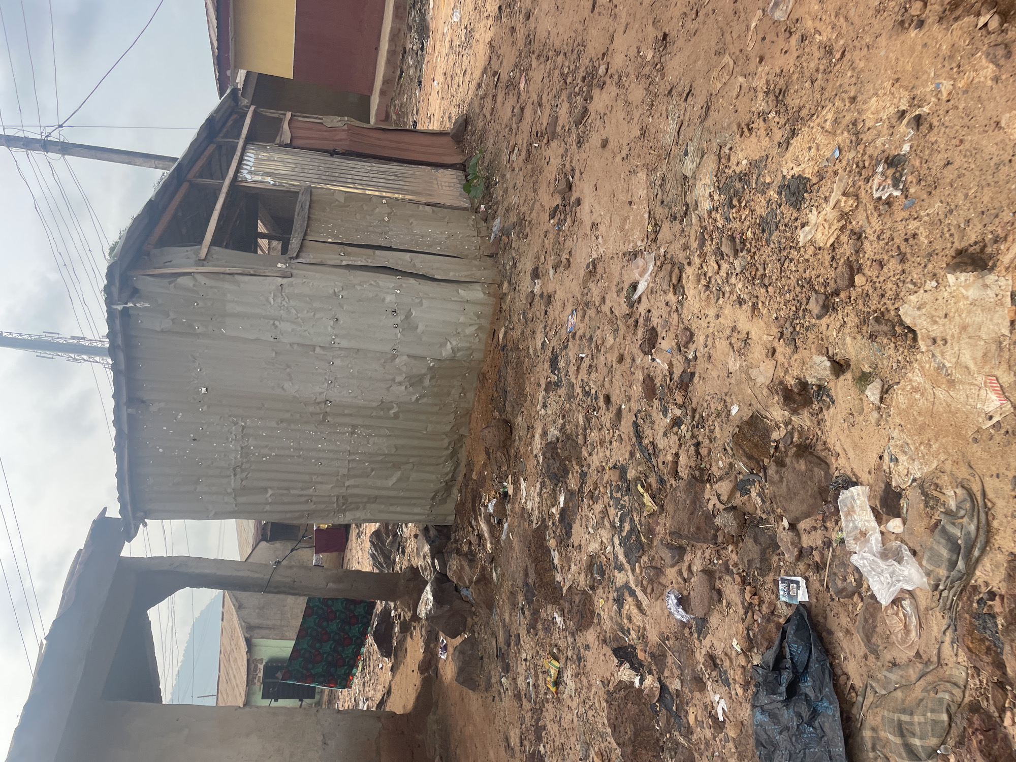 In a dusty and debris-strewn area in Lokoja, Nigeria, a makeshift structure constructed from corrugated metal stands on the right. The structure appears weathered and possibly unfinished, with exposed wooden beams supporting its roof and an open side. In the foreground, the ground is a mixture of reddish-brown dirt, rocks, and various pieces of refuse, including plastic bags, torn fabric, and other discarded items. To the left of the metal structure, another building with a simple roof is visible, and a colorful piece of fabric is draped over a support. Power lines crisscross the sky above, and in the background, other buildings and a distant hillside can be seen. The overall scene depicts a rugged and somewhat impoverished environment.