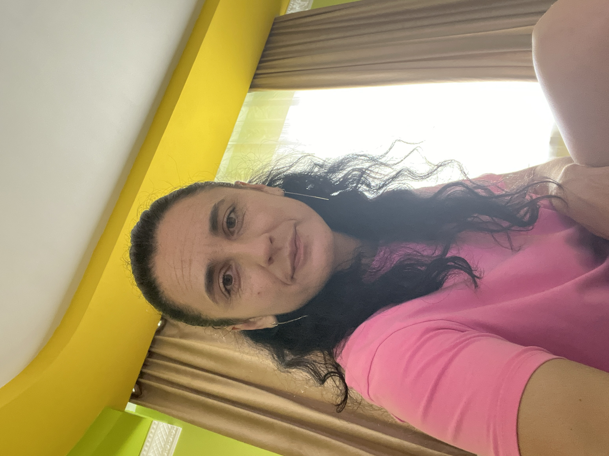 An indoor, slightly low-angle photograph taken in Onokivtsi, Ukraine, captures a woman with dark, wavy hair pulled back, wearing a short-sleeved pink t-shirt and thin, chain-like dangling earrings. She looks directly at the camera with a composed expression. The background features a room with a prominent bright yellow ceiling and a lime green wall section on the left, separated by white decorative trim. Brown curtains frame a large, overexposed window on the right, casting bright light into the room. The woman's bare leg is partially visible on the bottom right of the frame.