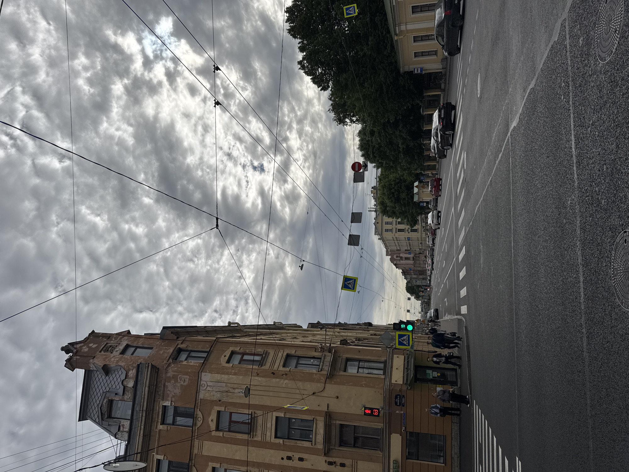 This is a photograph taken on a cloudy day in Saint Petersburg, Russia, on August 24, 2023, at approximately 16:30 local time. The scene captures a street view with a prominent building on the left, characterized by its light brown facade, multiple windows, and an ornate mansard roof. Several cars are visible on the street, including a black sedan and a dark-colored sedan in the foreground. Pedestrians are present on the sidewalk and crossing the street at a marked crosswalk. Traffic signals indicate a green light, and pedestrian crossing signs are displayed. Overhead, a complex network of wires crisscrosses the sky, suggesting the presence of public transportation like trams. The overall atmosphere is urban, with elements of historic architecture and modern city life.