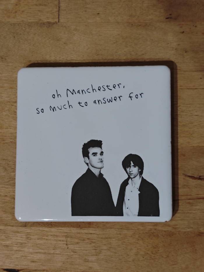 The image displays a white square coaster or tile resting on a light brown wooden surface. The wooden surface appears to be a tabletop or floor, characterized by visible grain patterns and horizontal seams between planks.
The coaster itself has rounded corners and a slightly raised edge, suggesting it might be made of ceramic or a similar hard material. The primary design on the coaster is a black and white illustration featuring text at the top and two figures below.
The text, written in a casual, somewhat handwritten script, reads:
"oh Manchester,
so much to answer for"
Below the text, two men are depicted in a black and white, somewhat stark, illustrative style. Both are shown from the chest up.
The man on the left has dark, slightly messy hair styled upwards at the front. He is looking directly at the viewer with a subtle, perhaps slightly defiant, expression. He is wearing a dark, high-collared shirt or jacket.
The man on the right has darker, shaggier hair that covers part of his forehead. He is also looking towards the viewer, though his gaze seems a bit more reserved or pensive. He is wearing a lighter-colored shirt, possibly white or light grey, with a dark jacket or blazer over it, and the collar of the shirt is visible.
The overall impression is that of a piece of merchandise or art related to the iconic British band The Smiths, given the famous lyric (from their song "Suffer Little Children") and the recognizable likenesses of lead singer Morrissey (left) and guitarist Johnny Marr (right). The image is well-lit, showing no harsh shadows, and the details of both the coaster and the wooden background are clear.