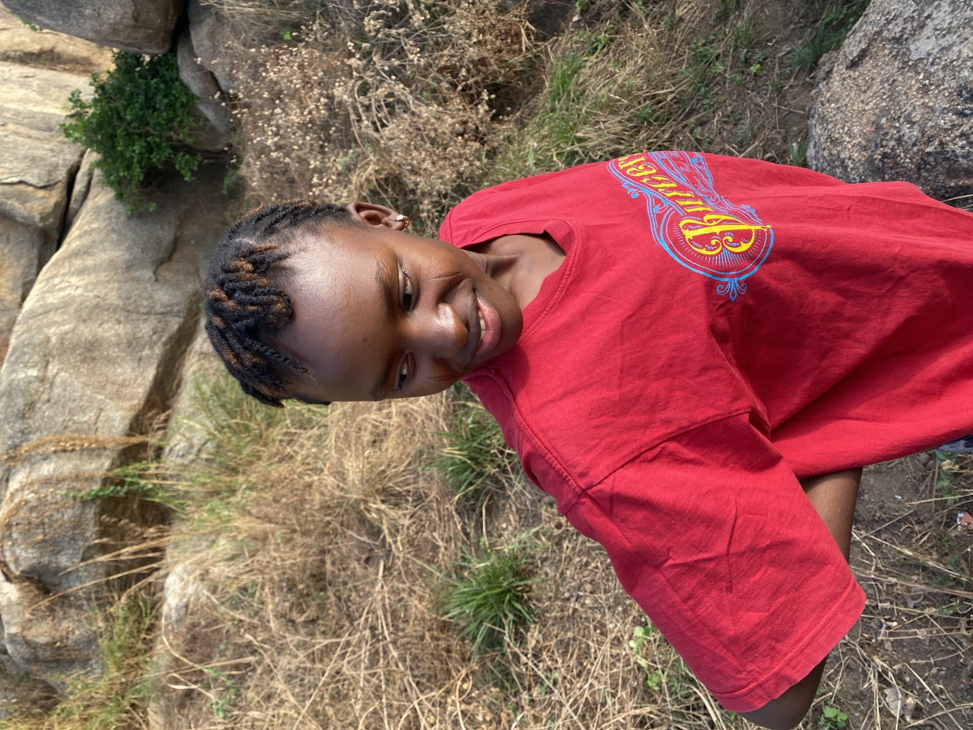 A young, dark-skinned woman with braided hair is smiling. She is wearing a red t-shirt with a graphic design on the upper back. The background indicates an outdoor location in a rural or semi-rural area, with large rocks and dry grass visible. The location is Vom, Nigeria.