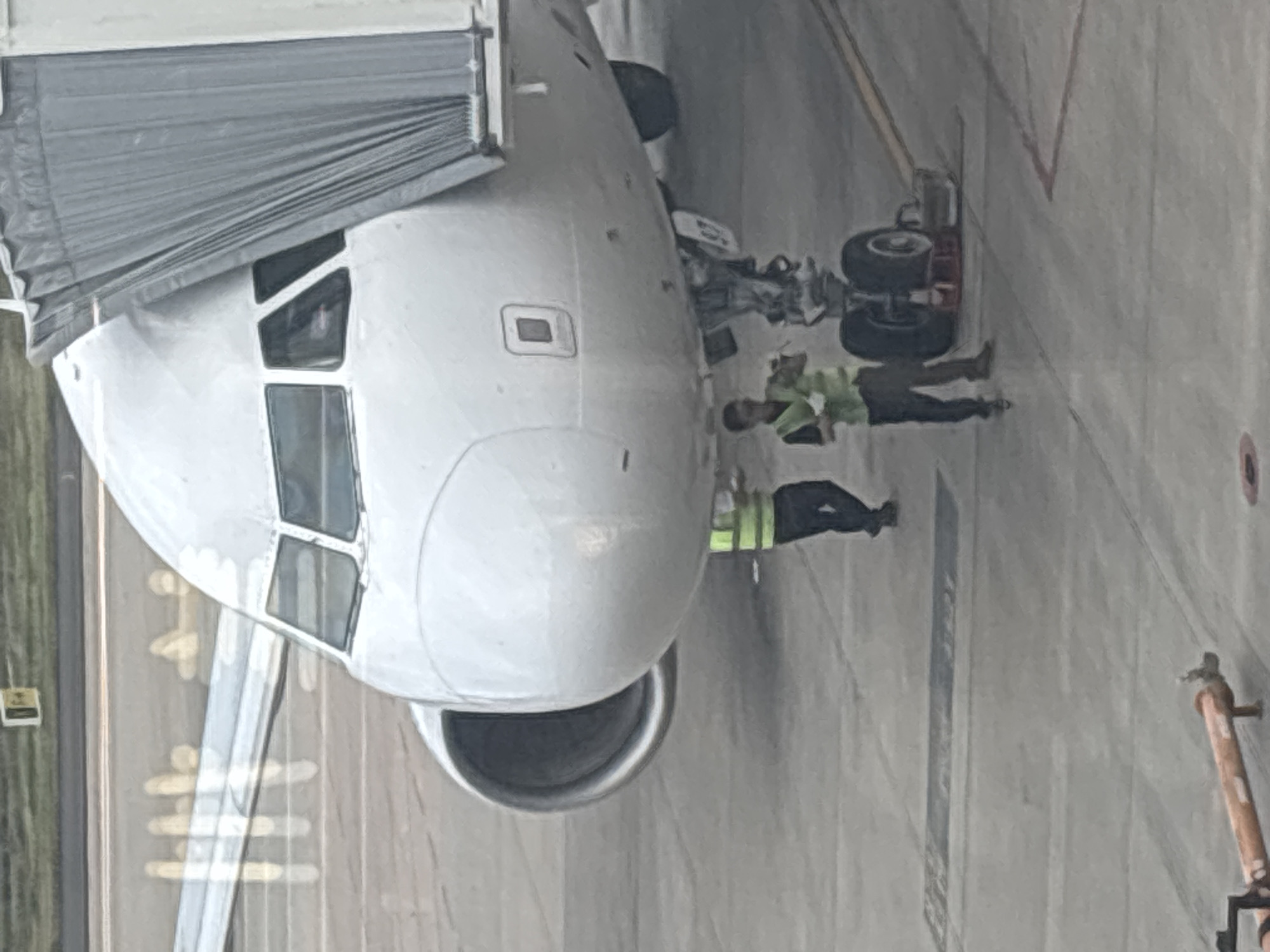 The image captures an outdoor scene at an airport tarmac, likely in Xinwu District, China, given the presence of an airplane and airport personnel. A large, white passenger airplane is the primary subject, with its front section, including the cockpit and engine nacelle, prominently displayed. The aircraft is connected to a jet bridge, indicated by the grey, accordion-like structure covering the passenger entry.
Two individuals, presumably airport ground crew, are visible near the airplane's landing gear. One person, wearing a bright yellow safety vest over dark clothing, is standing near the nose gear and appears to be holding some documents or equipment. Another person, also in a yellow vest and dark pants, is further back and to the left, near the main landing gear, and seems to be observing or directing. The tarmac is a concrete surface with visible markings and lines. In the background, there is a glimpse of a grassy area and what appears to be an airport runway or taxiway with painted lines. A piece of orange equipment, possibly a chocks or tow bar, is visible in the lower left corner of the image. The overall impression is one of a routine airport operation.