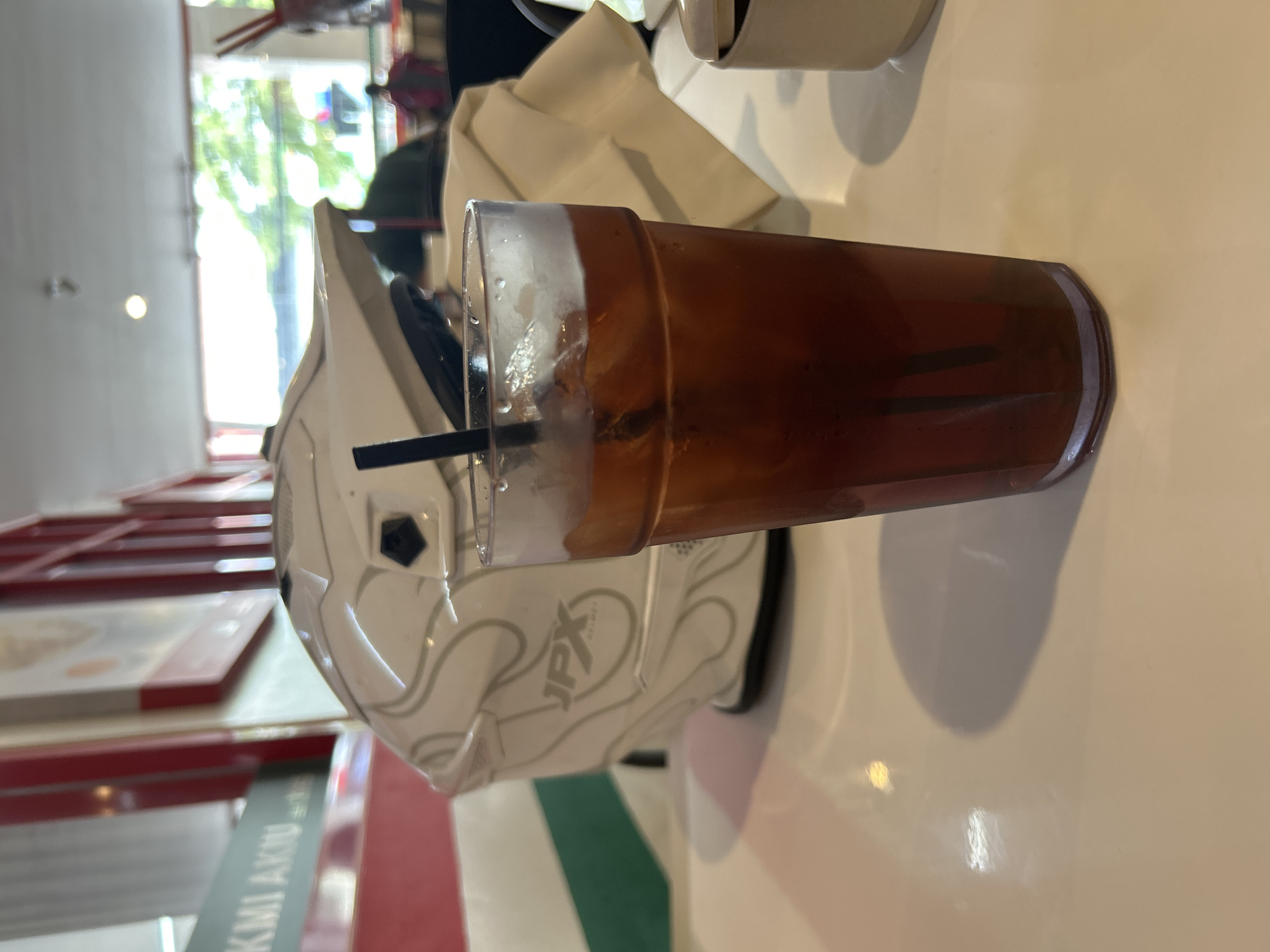 A glass of iced tea with a black straw sits on a white table, in front of a white motorcycle helmet with silver graphics. The helmet is labeled "JPX HELMET". In the background, there are red and white striped walls, a window with green foliage visible outside, and several people seated at tables. The setting appears to be an indoor dining area, possibly a cafe or restaurant, located in Denpasar, Indonesia. The lighting suggests it is daytime.