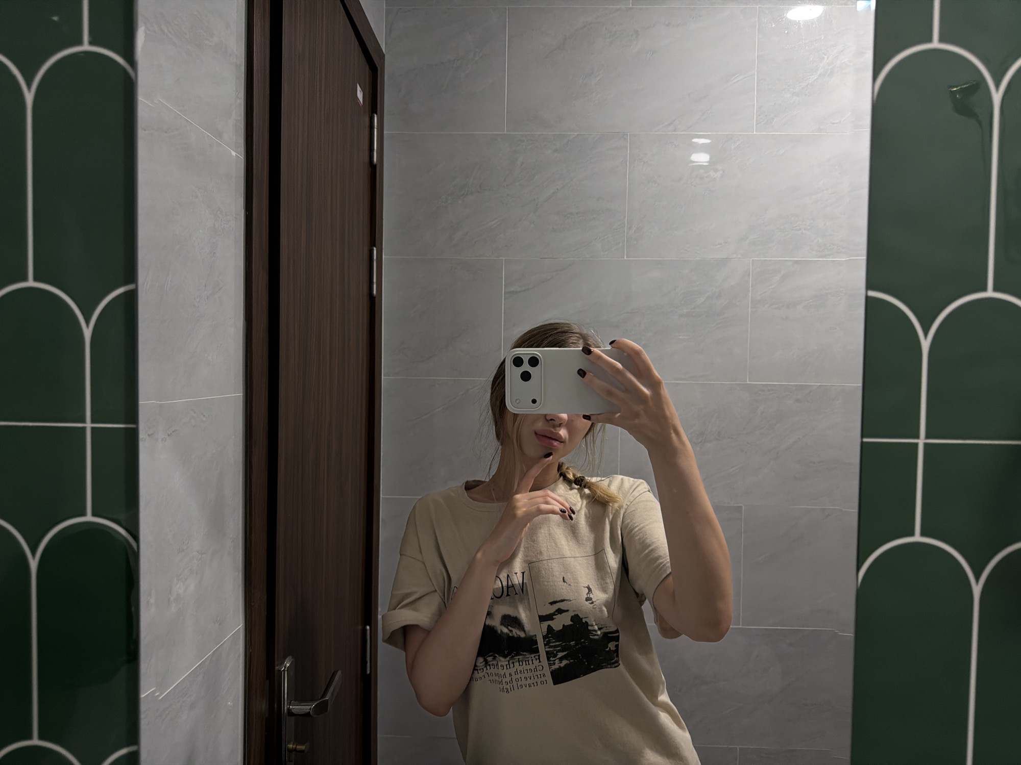 In a modern bathroom setting, a young woman with blonde hair braided to the side is taking a mirror selfie. She is wearing a light beige t-shirt with a graphic print. She is holding a white smartphone in her right hand and using her left hand to lightly touch her chin, creating a pensive pose. The bathroom features light grey marble-patterned tiles on the walls and dark wood doors. To the left and right of the central mirror are decorative green panels with white arch motifs. The scene is likely within a residential or hotel bathroom in Đà Nẵng, Vietnam, indicated by the architecture and interior design elements.