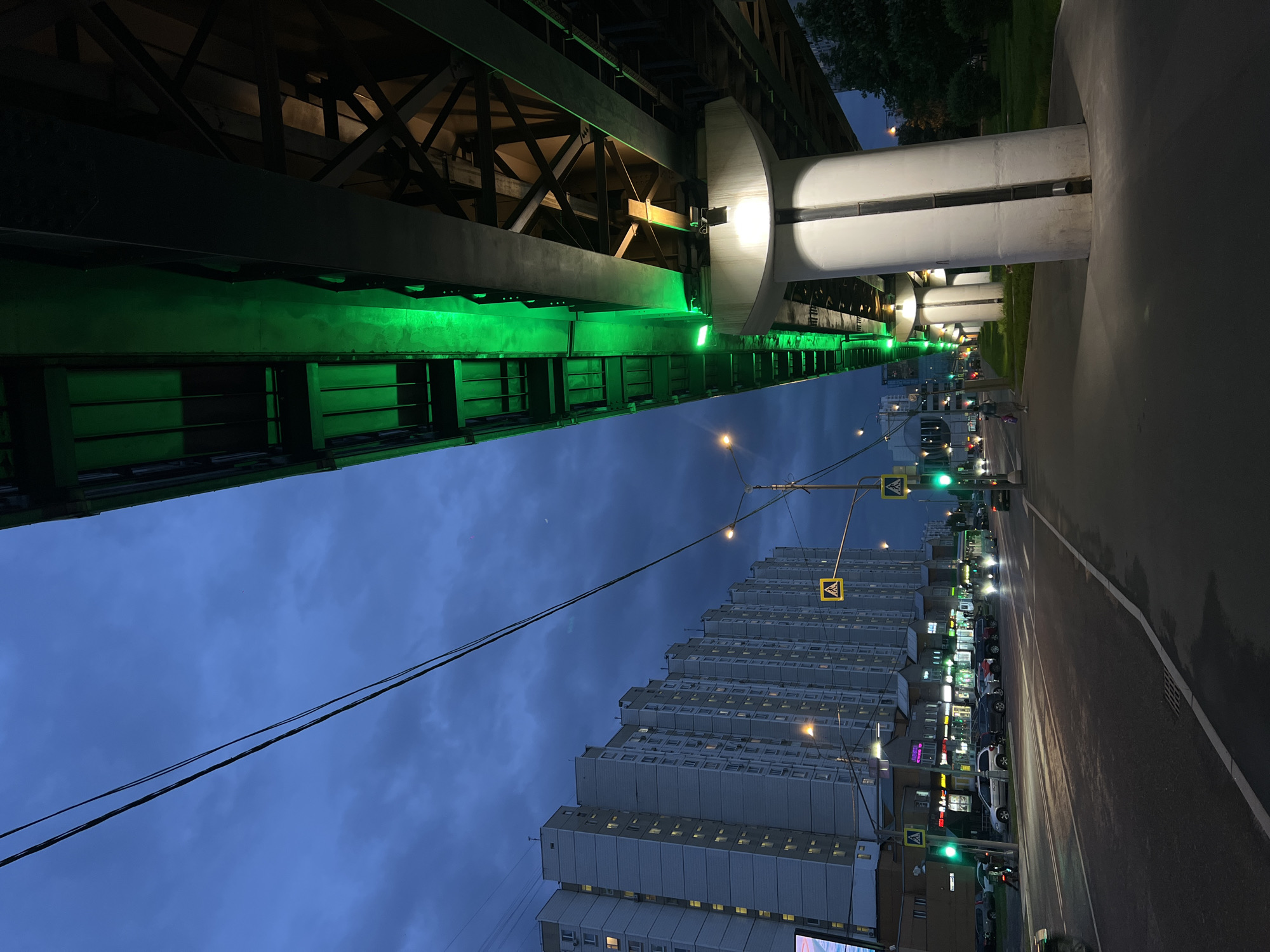 Captured at night, the image presents a Moscow, Russia cityscape. An elevated structure, possibly a bridge or overpass, dominates the left side, illuminated by green lights. Supporting columns, lit with white lights, stand along the right, adjacent to a road. The road is flanked by a high-rise building with many windows. Streetlights and lit signs are visible. Cars and other vehicles are parked along the road. The sky is a gradient of blue. Power lines stretch across the sky, intersecting with the building and the elevated structure. Pedestrian crossing signs are seen along the road.