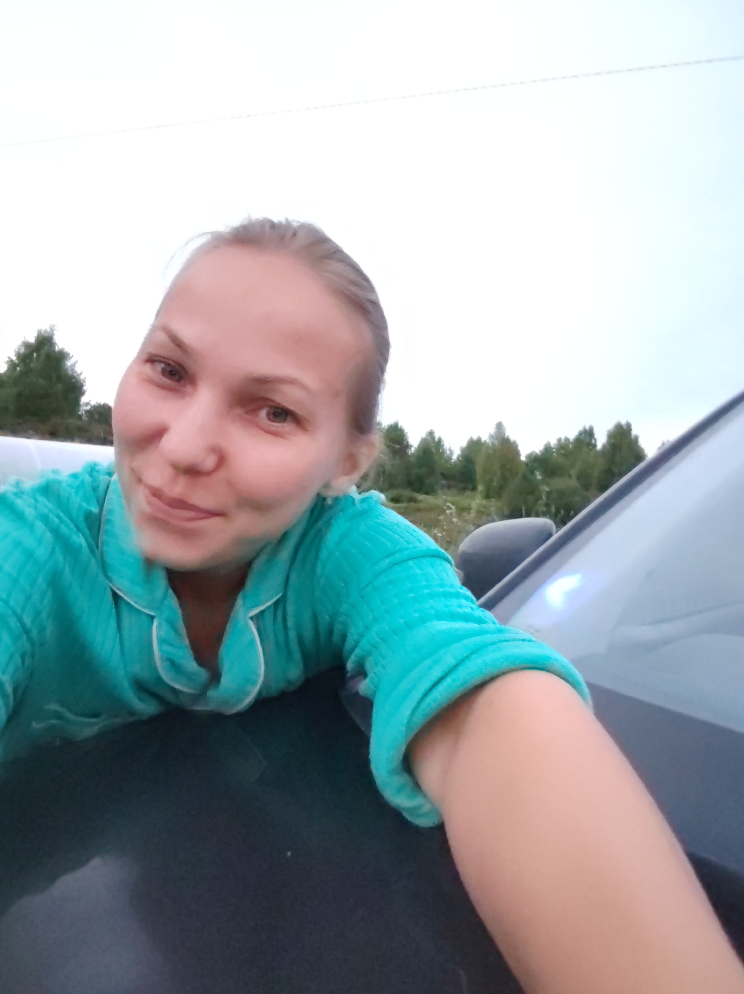 A woman is taking a selfie outdoors, leaning on the hood of a dark-colored car. She is wearing a teal pajama top with white trim. The background shows a slightly overcast sky and a line of trees. The location is indicated as the city of Chukhlem, Russia.