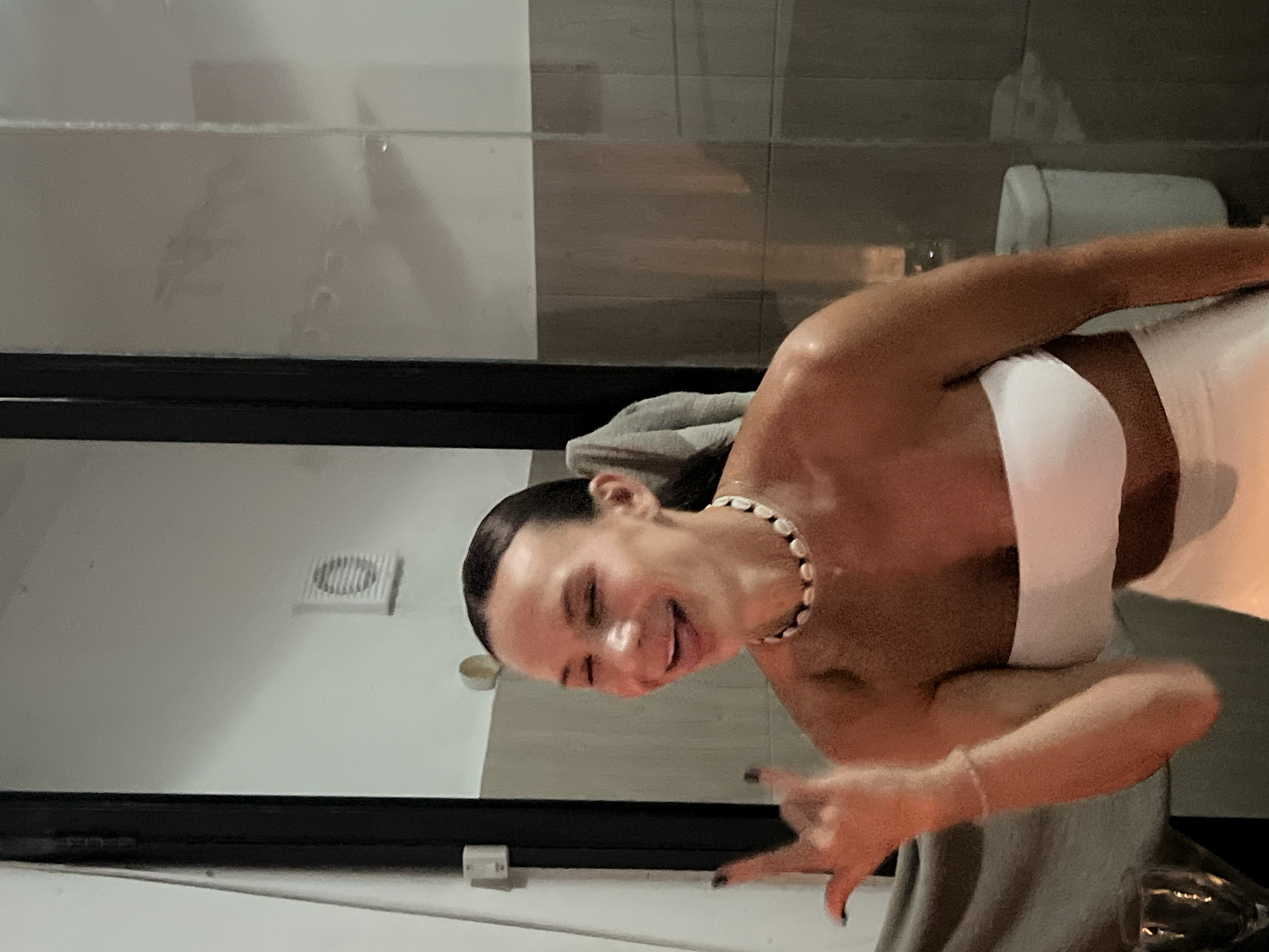 A young woman with her hair pulled back is pictured in a dimly lit bathroom, possibly in Unknown, Indonesia. She is wearing a white strapless top and white pants, with a shell necklace. Her eyes are closed, her tongue is sticking out, and her left hand is raised with fingers spread in a rock-and-roll gesture. The background includes tiled walls, a ventilation grate on the upper left, and a toilet visible to the right. A towel hangs on a glass door on the left side of the image, and a wine glass is partially visible in the lower left corner. The lighting suggests an artificial light source.