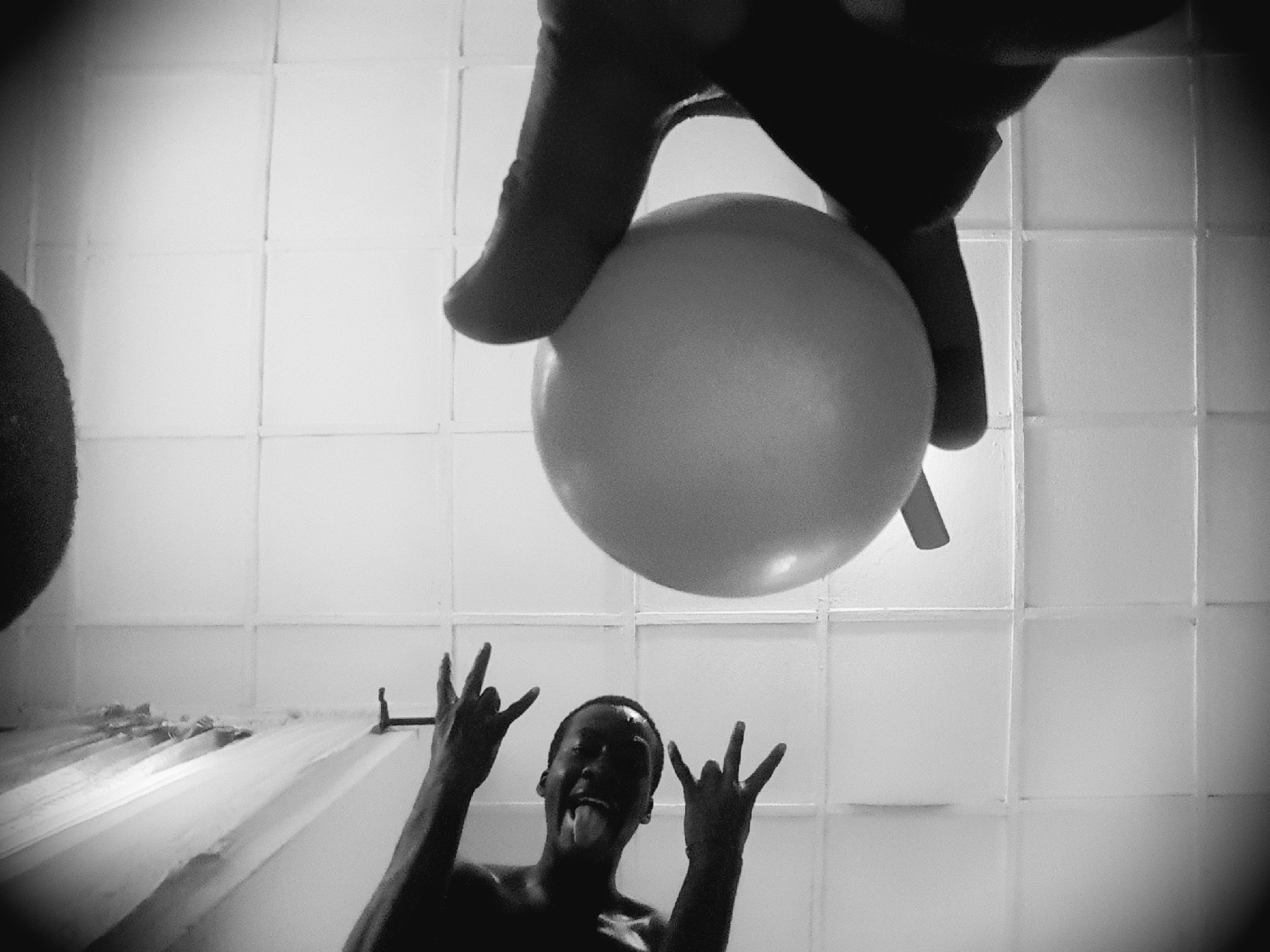 An extreme low-angle, black and white photograph shows a large, pale, spherical object, likely a balloon, held by a dark, silhouetted hand and thumb in the lower left of the frame. The object is positioned in the foreground and slightly to the left of the center. In the middle ground and to the right, a person, also silhouetted and appearing to be submerged or partially wet, is visible from the chest up. Their head is tilted back, mouth open, and their face is glistening. Both of their hands are raised, with fingers extended in a gesture resembling the "rock on" sign. The background consists of a tiled wall, likely in a bathroom, with a shower curtain visible on the far right. The lighting is harsh, creating strong shadows. The overall impression is of a playful or dramatic moment captured from an unusual perspective.