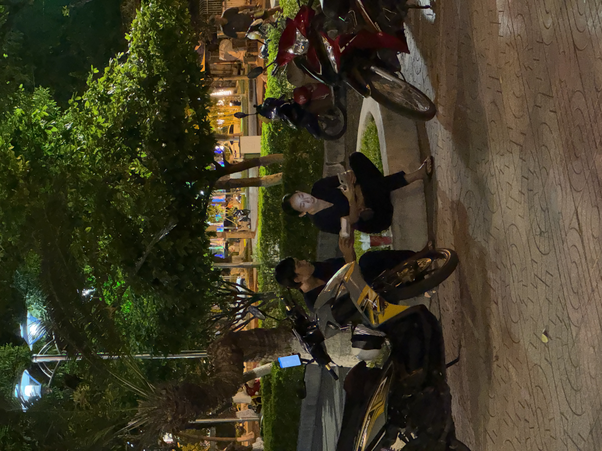 Two individuals are seated near parked motorcycles on a stone-tiled pathway at night. The scene is dimly lit, suggesting it's nighttime. One person, wearing black clothing, is seated on a low concrete structure, looking at their phone. The other person, also in black, is leaning back and looking to the side. Several motorcycles are parked nearby, with one in the foreground displaying yellow and black accents. Lush green trees and foliage are visible to the left of the pathway, with some lights peeking through the leaves. In the background, there are hints of buildings and more lights, suggesting an urban or semi-urban environment. The overall mood appears relaxed, with the individuals engaged in their own activities. The time of day is clearly night, and the weather appears to be clear. There is no visible text in the image.