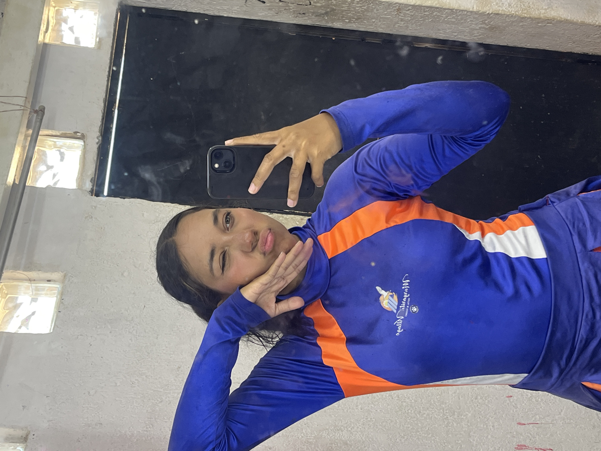 A young woman is taking a selfie in front of a dark mirror. The woman wears a blue long-sleeved athletic shirt with orange and white accents and is holding a black smartphone. She is making a kissing face and winking. The environment includes a weathered, light-colored wall with small window openings and a dark mirror surface. The location is likely indoors, possibly a changing room or bathroom, but the specific location in Porlamar, Venezuela, cannot be determined from the visual information alone.