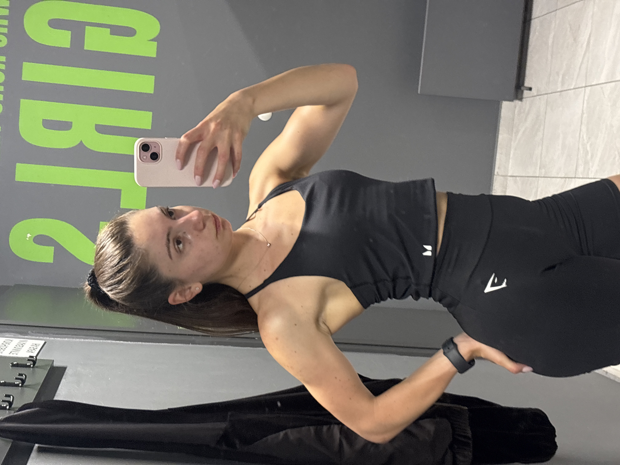 The image depicts a woman in athletic wear taking a mirror selfie. She is wearing a black sports bra and black high-waisted shorts with white logos. Her hair is pulled back in a high ponytail. She is holding a light pink smartphone in her right hand, which is angled to capture her reflection. A black smartwatch is visible on her left wrist.
The background suggests a gym or fitness facility. A dark jacket hangs on a rack to the left. To the right, large, bright green lettering is visible on a gray wall. The lettering appears to spell "GIRLS" backwards, with the "G" and "I" prominently displayed. The floor is tiled with a light gray pattern. The overall impression is a typical locker room or workout area. The context of Varna, Bulgaria is not directly evident from the image itself, but if this location is specified, it would be understood to be within a gym in that city.