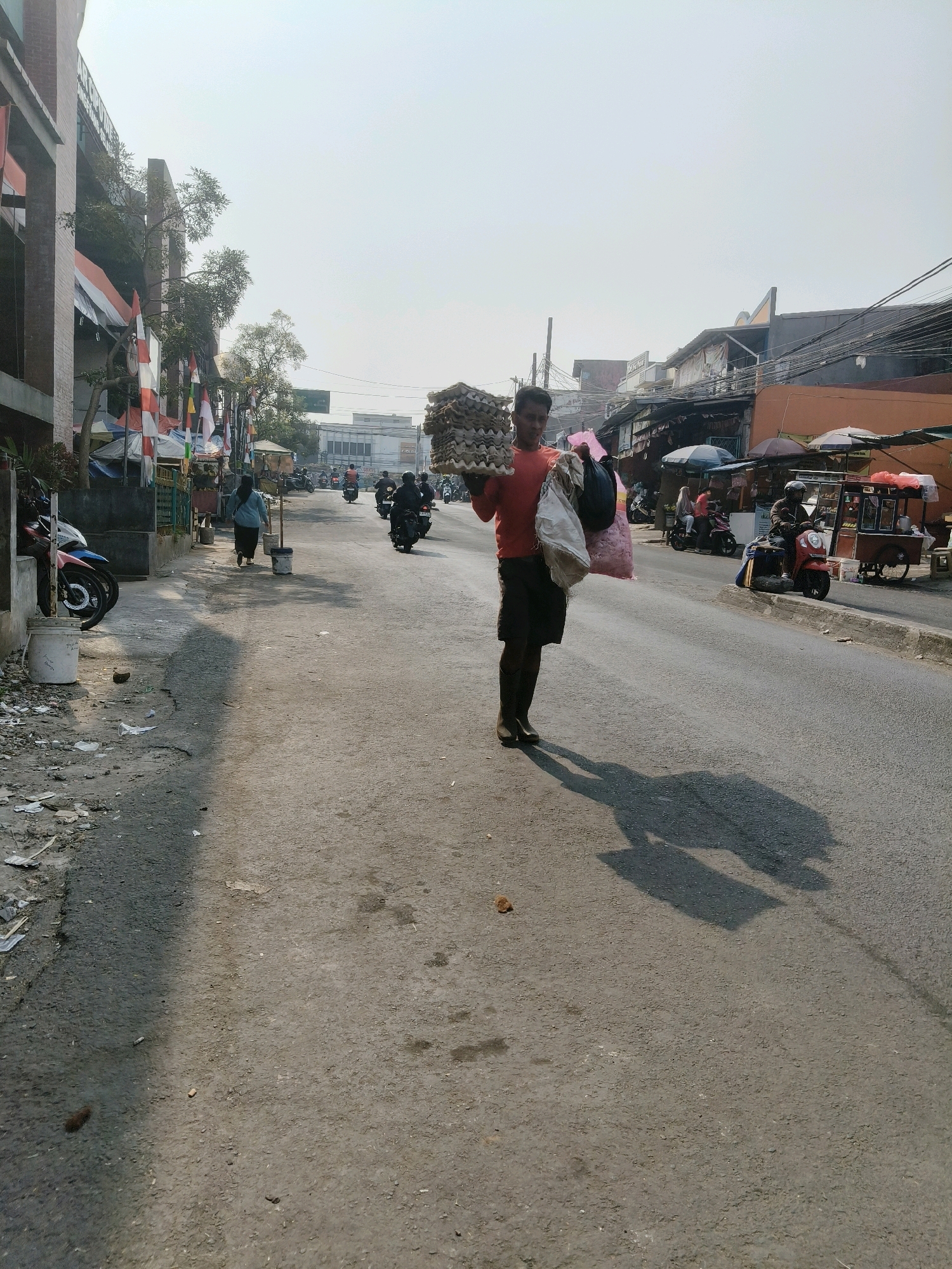 The image captures a street scene in South Tangerang, Indonesia, on a sunny day. In the foreground, a man wearing an orange t-shirt and dark shorts carries a stack of egg cartons in his left arm and two large bags in his right arm. He is walking on a paved road, casting a long shadow. The road is bordered by buildings and shops on both sides, with motorcycles parked along the left side. Several people are visible in the background, including a woman walking away from the camera and individuals riding motorcycles and standing near market stalls. The environment appears to be a bustling urban area with typical street infrastructure, including power lines and various vendor carts. The sky is clear, indicating fair weather.