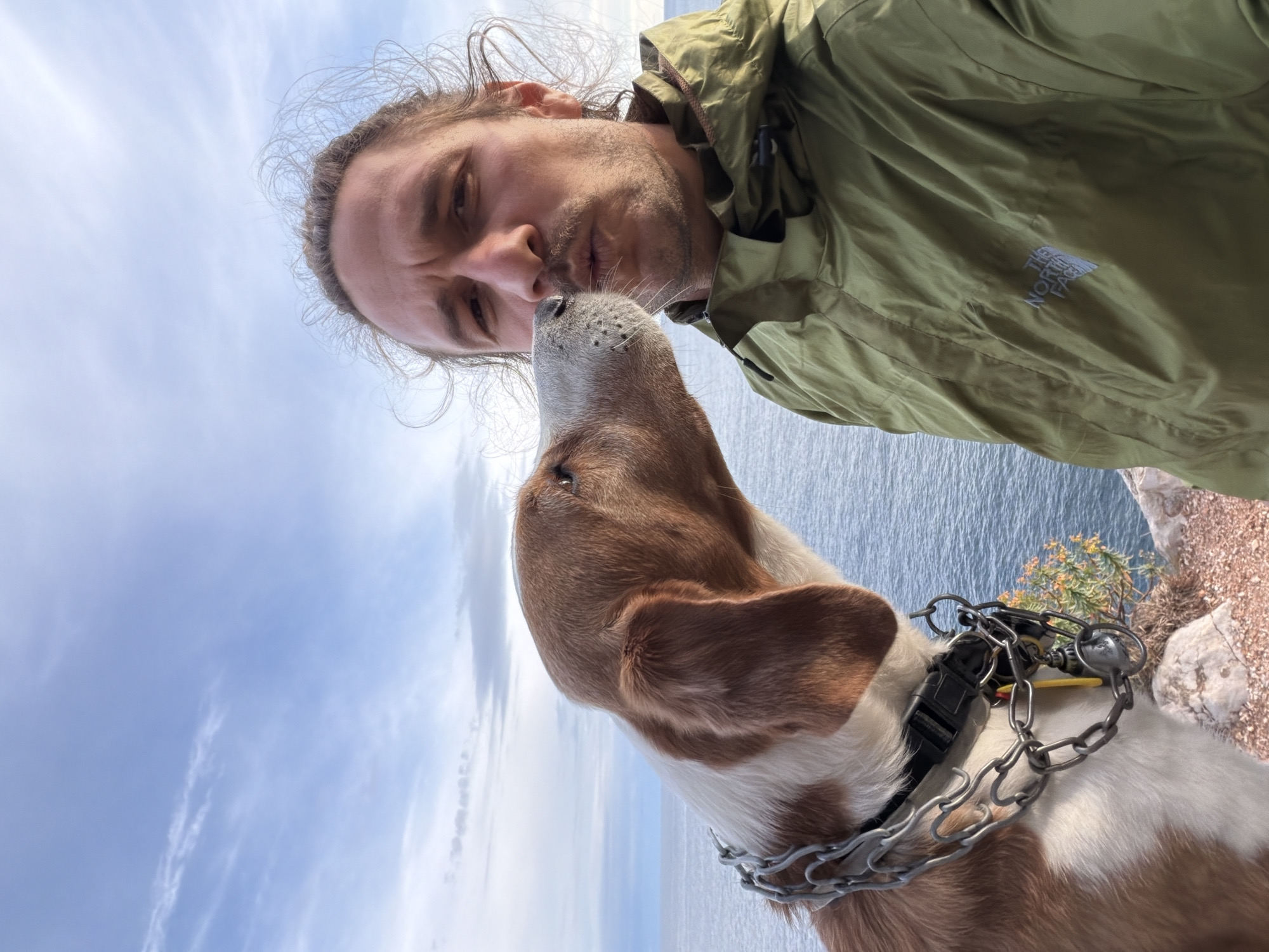 A selfie shows a man and a dog on a hill at Mirište, Montenegro, with the sea and sky in the background. The man, wearing a green jacket, is looking down at the dog, their noses nearly touching. The dog, with a brown and white coat and a metal chain collar, is looking up at the man. In the background, the sea extends to the horizon with the sky visible above. Some vegetation and rocks are visible on the hillside.