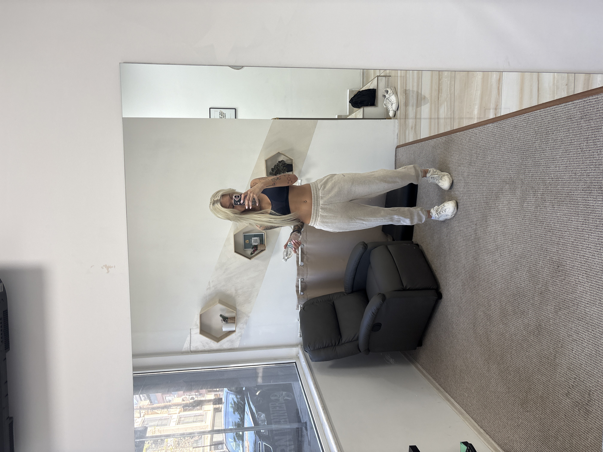 A young woman with long, straight blonde hair and visible arm tattoos is captured in a mirror selfie. She wears a dark sports bra, light-colored sweatpants, and white sneakers, with a belly button piercing visible. She holds a smartphone up to take the picture, and a plastic water bottle in her other hand.
The setting appears to be an indoor room, possibly a waiting area or lounge, within a commercial building in Plovdiv, Bulgaria. A large full-length mirror, in which the woman is reflected, dominates the central part of the image. To her right, a dark grey recliner chair is placed on a light grey textured carpet. The wall behind the chair is painted in a combination of white and a diagonal light grey/beige stripe, featuring two hexagonal wooden shelves, each holding small decorative items or plants.
Reflected in the mirror, on a shelf or ledge, are a pair of white sneakers and a black bag or piece of clothing. A framed poster with the text "Don't wish for it work for it" is also reflected on the wall above. Through a large window on the left, an urban street scene is visible, including parts of buildings and a car with the word "PANTHER" partially legible on its side. Natural light suggests it is daytime.