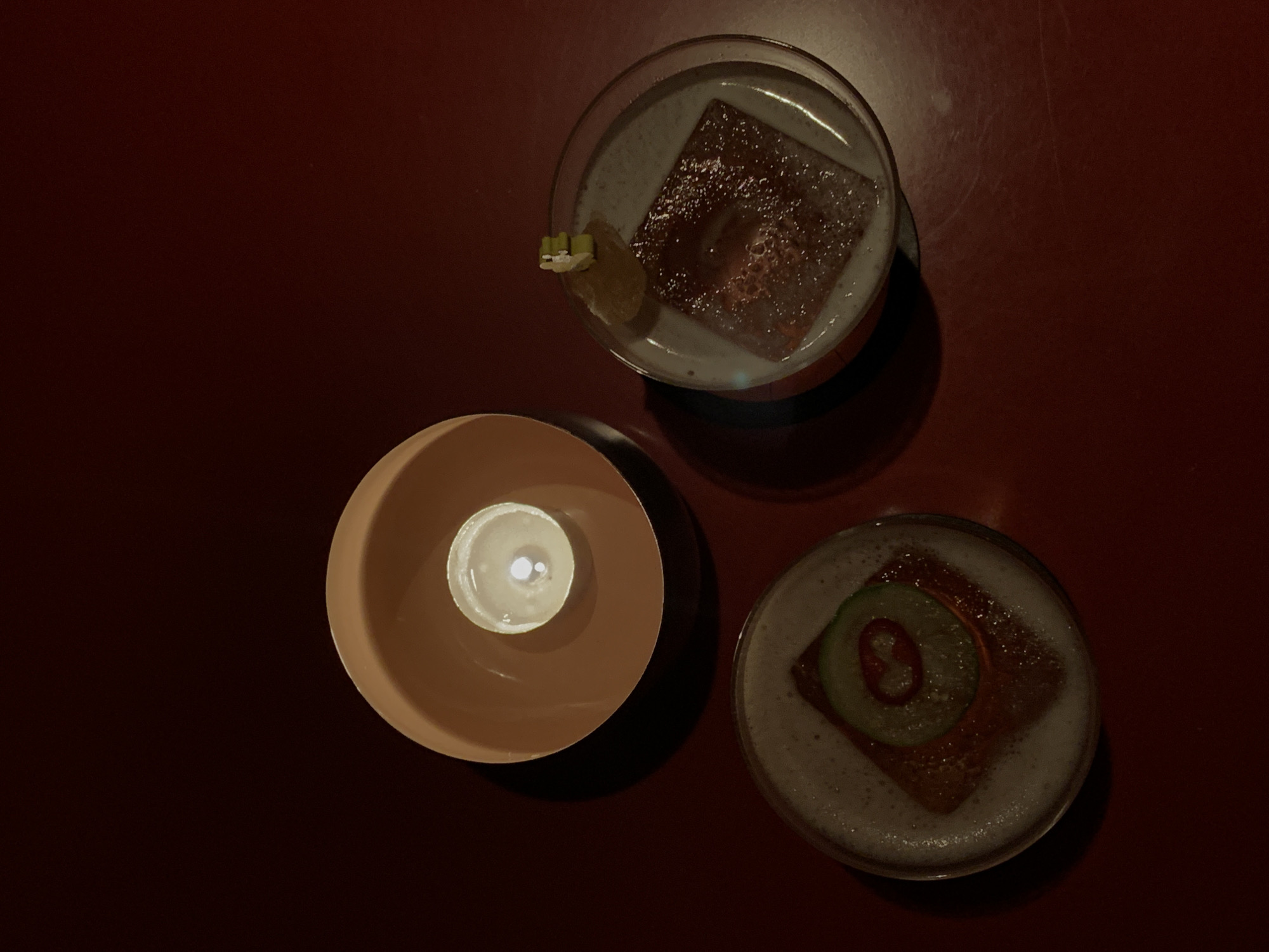This overhead shot depicts two artfully prepared cocktails and a lit candle arranged on a dark, reddish-brown table, likely within a dimly lit bar or lounge in London, United Kingdom. The low lighting suggests an evening or nighttime setting, creating a moody and intimate atmosphere.
One cocktail, positioned in the upper central area, features a frothy, light-colored head over a dark, reddish liquid. A large, square ice cube is visible within the drink, and it is garnished with a small lime wedge and a few tiny green cubes on the rim of the glass.
The second cocktail, located in the lower right, also presents a frothy top layer above a dark liquid. Its distinctive garnish includes a thin, round slice of cucumber, with a smaller, bright red chili ring placed inside the cucumber.
Between the two drinks, a small candle burns brightly, contained within a beige, cup-shaped holder. The candle flame casts a soft, warm glow, highlighting the central elements of the composition.
The scene implies an activity of socialising and enjoying beverages. No individuals are visible in the frame, and there is no discernible text. The overall aesthetic is sophisticated and inviting, typical of a modern cocktail bar.