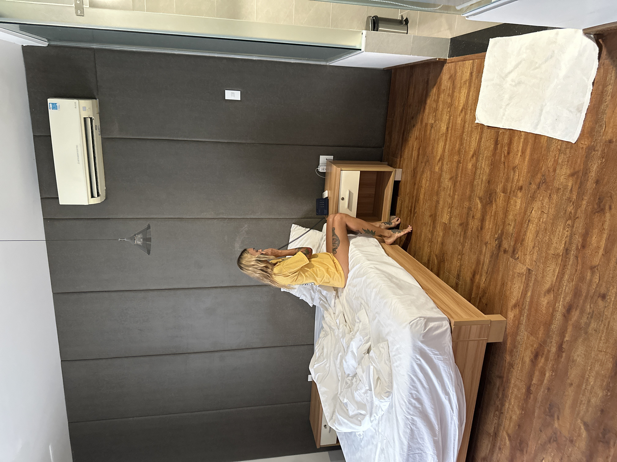 The image shows the interior of a hotel room, likely in Đồng Nai, Vietnam. The room has a bed with white linens, on which a woman with blonde hair is lying, talking on a cell phone. She is wearing a yellow shirt. The room has wood flooring and a dark grey upholstered wall. There is an air conditioning unit mounted on the wall, a bedside table, and a white rug on the floor.