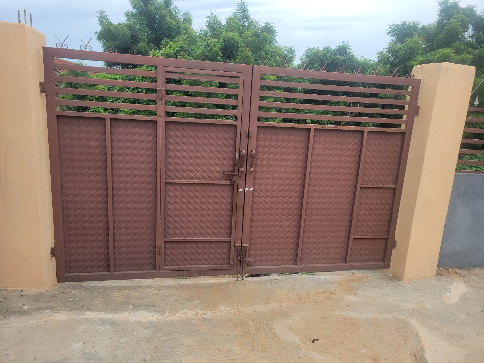 A pair of brown metal gates stands closed in front of lush green trees under a cloudy sky. The gates are constructed with horizontal bars at the top and a patterned metal filling in the lower portion. Decorative metal swirls are attached to the top edge of the gates. Each gate has a handle and a latch mechanism, suggesting they are secured.
The gates are flanked by two light-colored pillars, one on the left and one on the right. The pillar on the left is partially visible, showing a textured surface. The pillar on the right is more prominent, with a flat face and a rounded edge where it meets the gate.
The ground in front of the gates is a rough, concrete surface with some scattered stones and dirt. Behind the gates, dense green foliage of trees creates a natural barrier. The sky above is overcast, indicating a possibly humid or rainy day. The scene appears to be an entrance to a property, possibly a home or a compound, given the presence of the sturdy gates. There is no visible text in the image. The time of day is difficult to determine precisely due to the cloud cover, but it appears to be daytime. The overall atmosphere is neutral, with no strong emotional cues present.