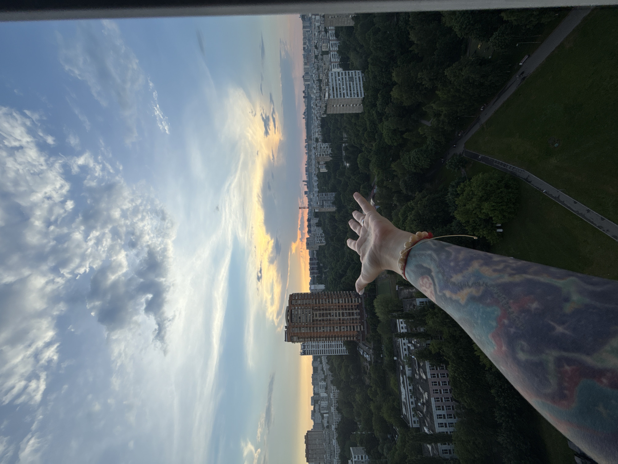 An overhead, wide shot captures a view of a city skyline at dusk, seen from an elevated position, likely a balcony or window. In the foreground, a person's left arm, heavily tattooed with a colorful, abstract design, extends into the frame, hand open as if reaching towards the sky. A bracelet of light-colored beads adorns the wrist.
The city below is densely populated with numerous apartment buildings and residential blocks, interspersed with a significant amount of lush green trees, suggesting a park or wooded area within the urban landscape. One prominent, tall apartment building with a reddish-orange facade stands out among the others. The sky above is a gradient of blues and whites, with scattered clouds tinged with the warm hues of the setting sun, creating a dramatic and vibrant sky. A sliver of dark vertical frame is visible on the right edge of the image, indicating the edge of the structure from which the photo was taken. The scene appears to be set in Moscow, Russia, given the architectural style and the presence of significant green spaces within a major city.