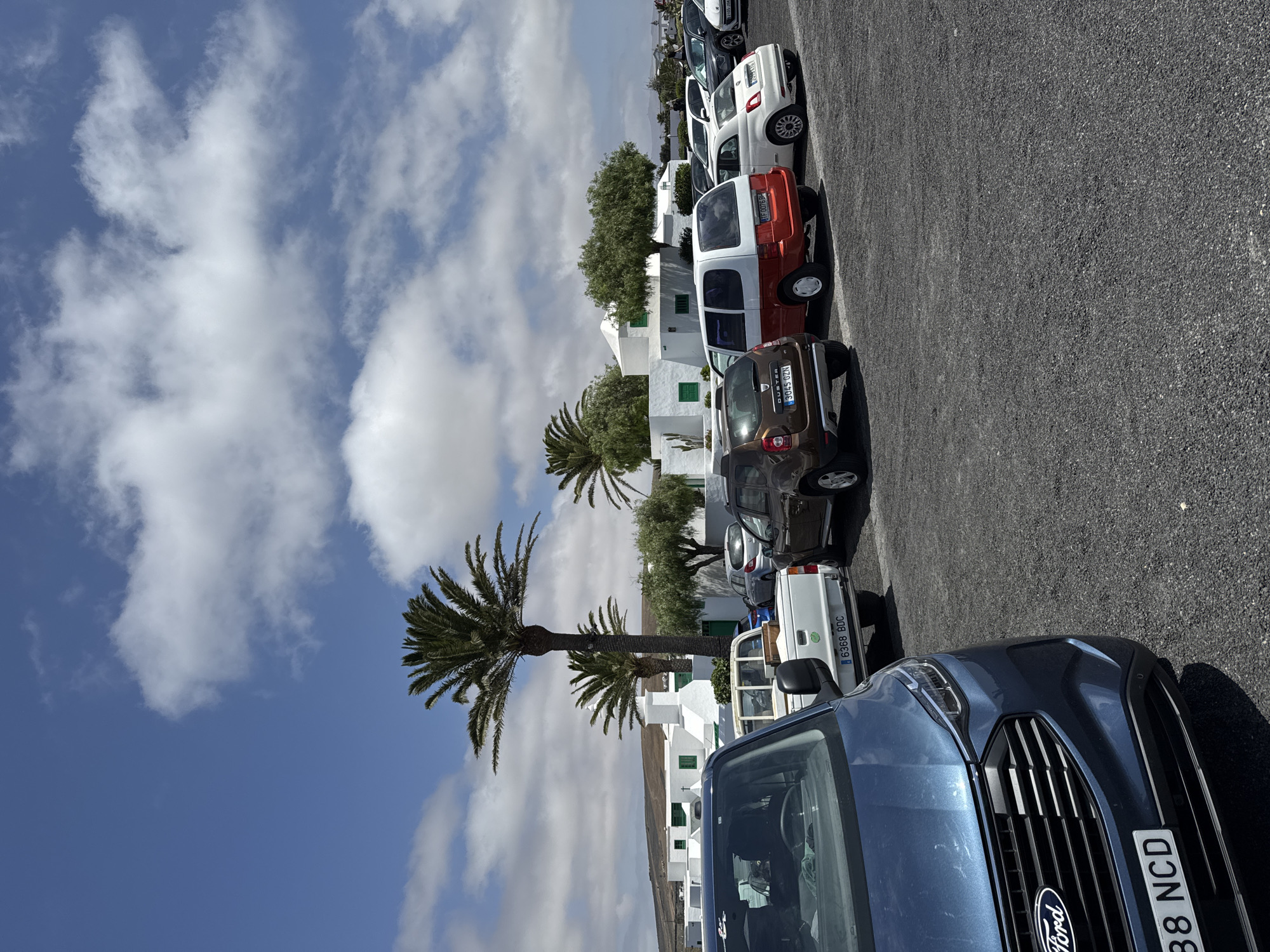 A parking lot scene in San Bartolomé, Spain, is captured under a partly cloudy sky. A line of parked cars fills the frame, with a blue Ford prominently in the foreground, displaying the license plate "4388 NCD". Other cars parked behind it include a white pickup with the plate "6368 BDC", a brown Renault Duster with "3645 GZN" on its license plate, and several other vehicles. The architecture in the background features whitewashed buildings with green window accents, typical of the region. Palm trees are interspersed amongst the buildings. The parking surface is paved with what appears to be dark asphalt or small gravel. No people are visibly interacting within the scene.