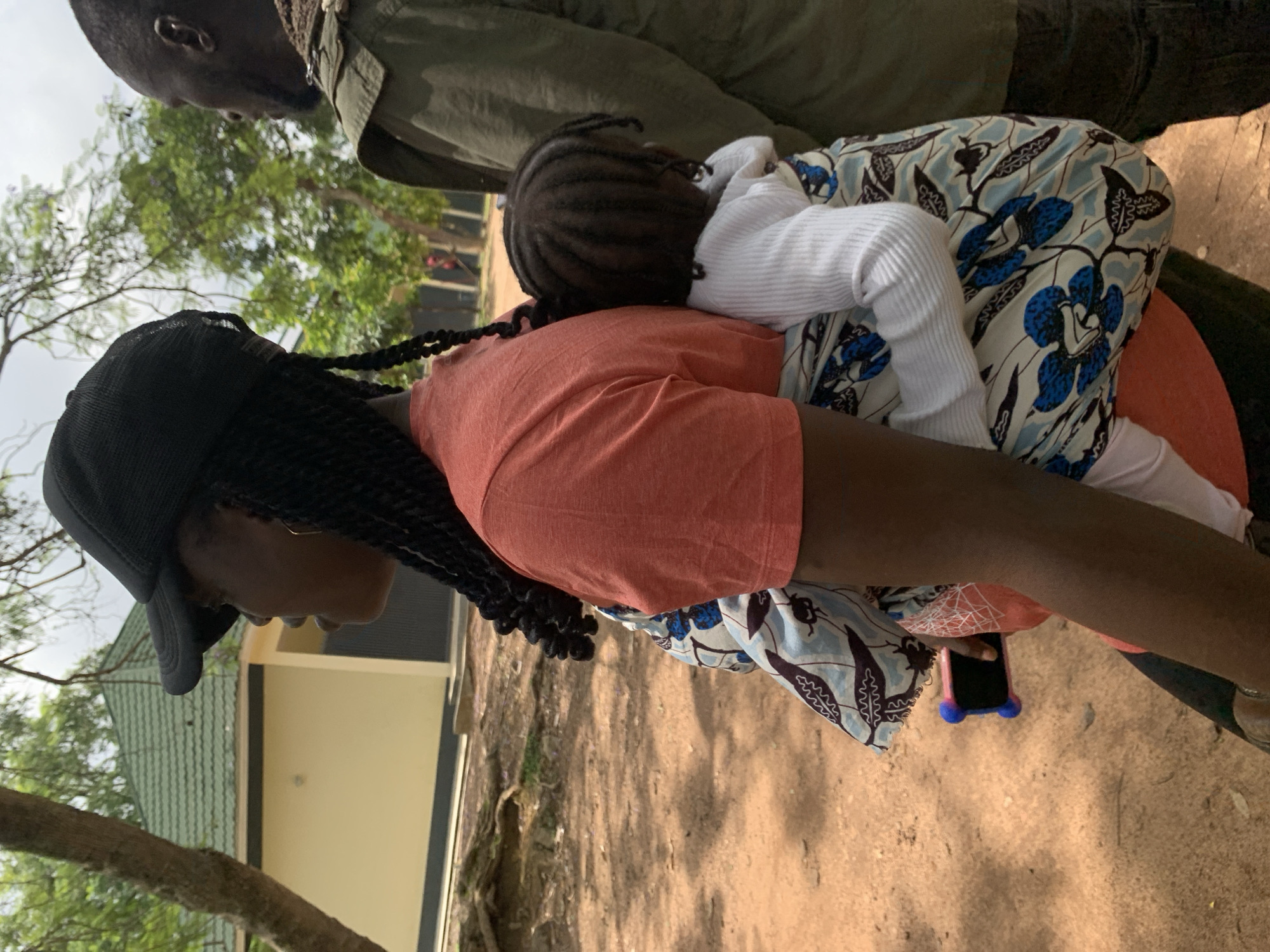 The media file shows a close-up, sideways view of two individuals, presumably a mother and child, in an outdoor setting. The foreground features the back of an adult wearing a coral-colored t-shirt and a black baseball cap. Long, dark braids cascade down their back. They are carrying a baby on their back, who is wearing a white long-sleeved shirt. The baby's head, with braided hair, is visible resting on the adult's shoulder. The baby is wrapped in a patterned cloth with blue floral designs and black leaf motifs.
In the background, a light-colored building with a green tiled roof is partially visible, suggesting a semi-urban or rural environment. Trees with green foliage and a dirt ground cover the rest of the scene. The lighting appears to be natural, indicating daytime, and the weather seems to be fair, with no visible rain.
A small, dark-colored smartphone with a pink case and blue protective bumpers is tucked into the adult's waistband, partially obscured by the patterned cloth. The overall impression is one of intimacy and care, with the adult carrying the child, likely engaged in a daily activity. The setting in Jos, Nigeria, provides a geographical context. No visible text is present in the image.