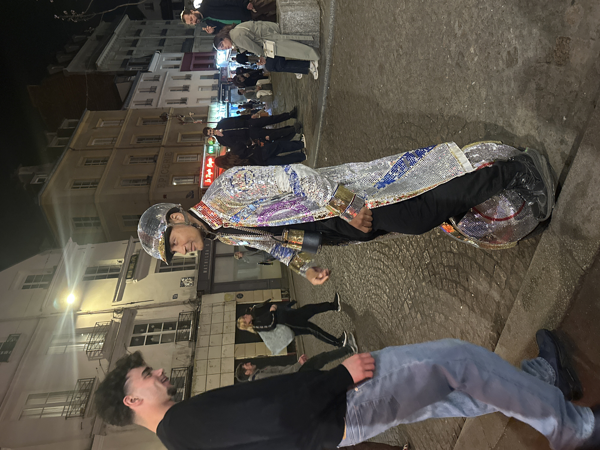 A night-time street scene in Paris, France, features a man dressed in a disco-ball suit and helmet. He stands on a large disco ball. The buildings are light-colored with multiple windows. Several people are walking along the street. A man in a black sweater and blue jeans is in the foreground. There are various shops and restaurants in the background, identifiable by their signs and awnings. One building reads "EPICERIE." Another says "Le Regale" in neon.