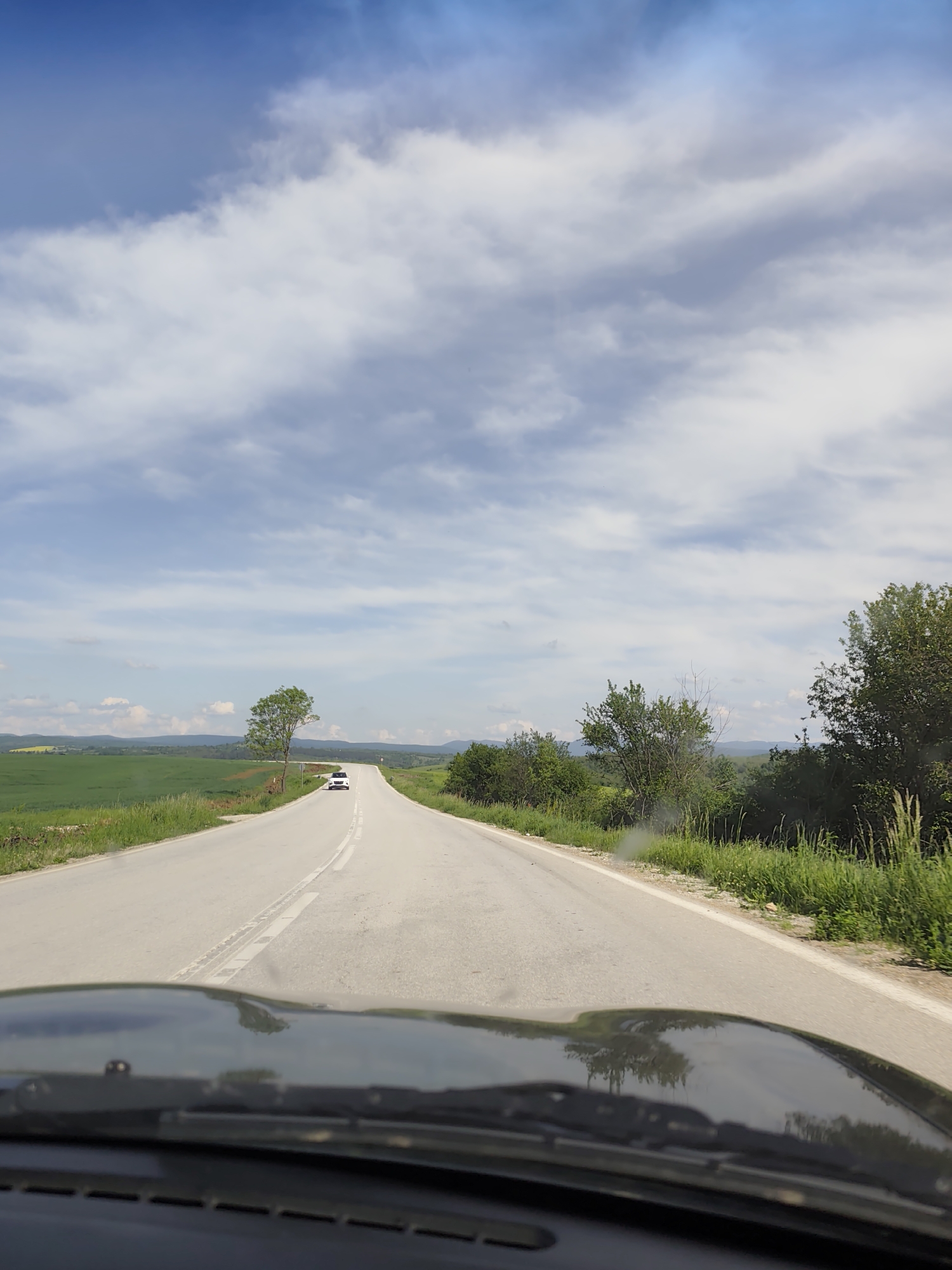 The image shows a view from inside a car, looking out onto a long, straight, asphalt road. A white car is visible in the distance on the road. The surrounding landscape includes green fields, trees, and bushes on either side of the road. The sky is blue with scattered clouds. The dashboard of the car is visible in the foreground.