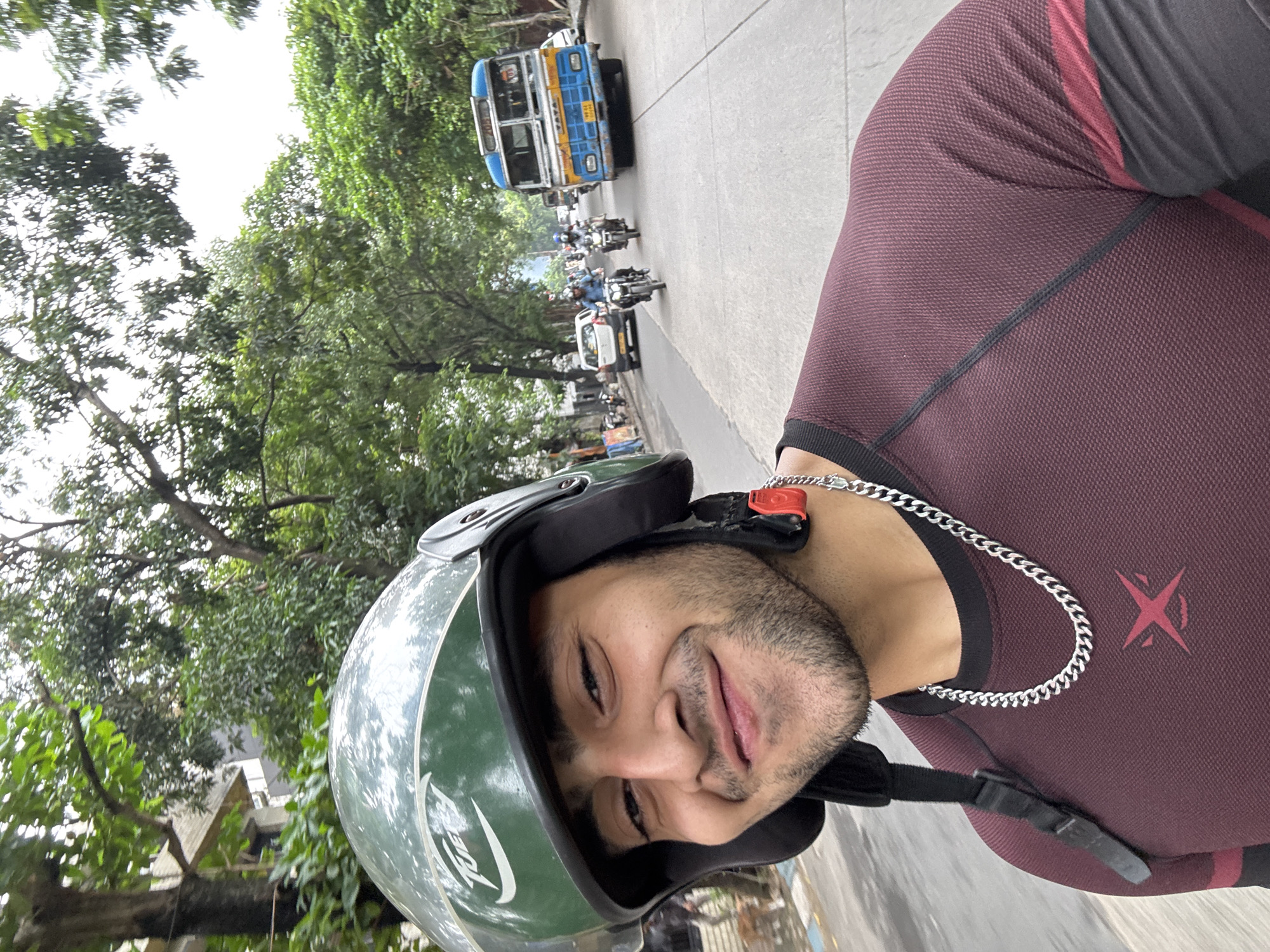 The image shows a man wearing a green helmet and a maroon t-shirt, with a silver chain around his neck. He is riding a motorcycle on a street in Kolkata, India. In the background, there are trees, buildings, cars, and a bus. The bus is blue and white and has "TATA" written on the front. The street appears to be busy with traffic. The man is looking at the camera and smiling.